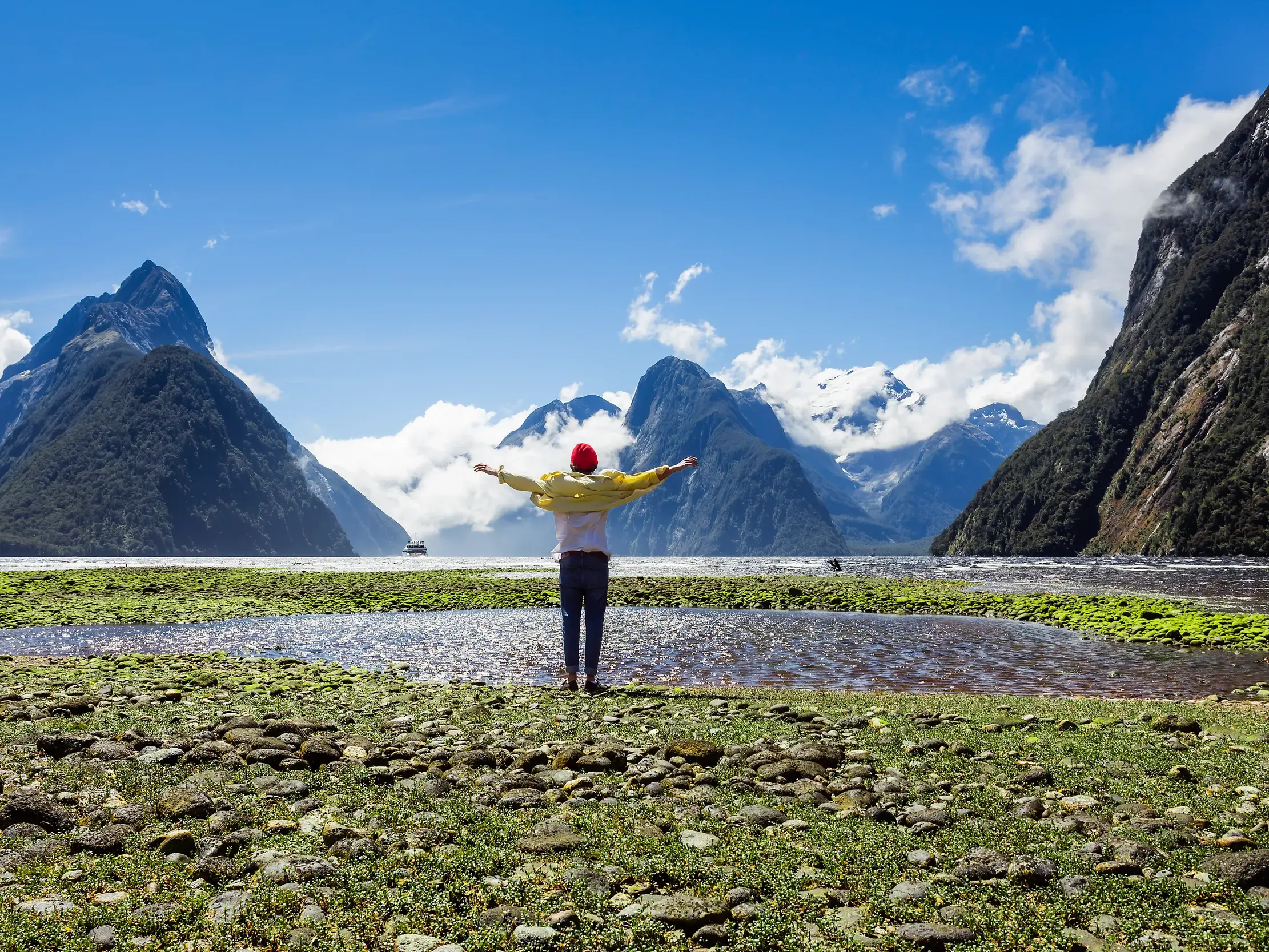 Eine Person mit ausgestreckten Armen blickt auf den Milford Sound mit Mitre Peak im Fjordland-Nationalpark, Neuseeland.