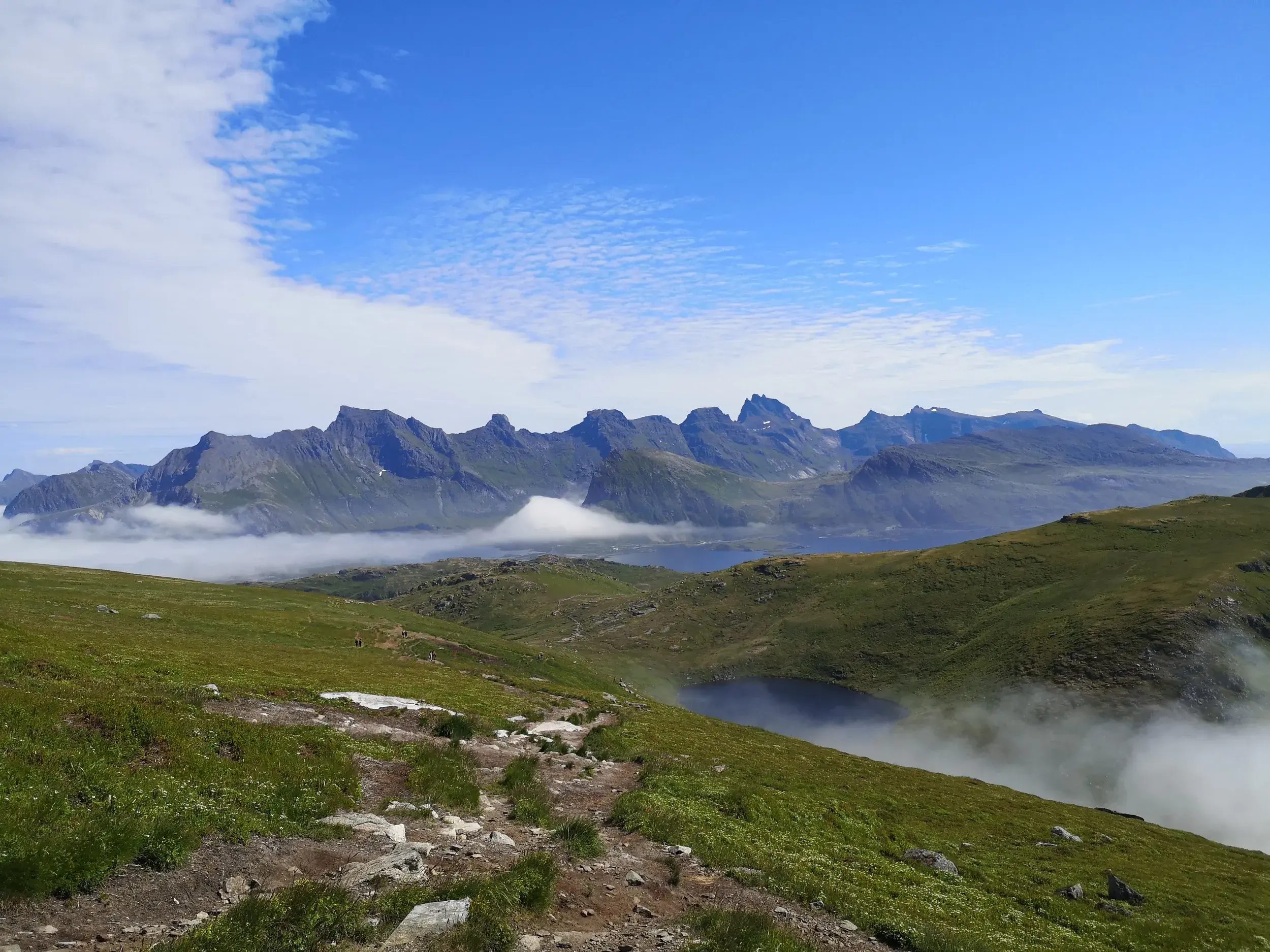 Panoramablick beim Wandern auf den Lofoten über einen Bergsee und Nebel hinweg zu einer schroffen Gebirgskette am Fjord.