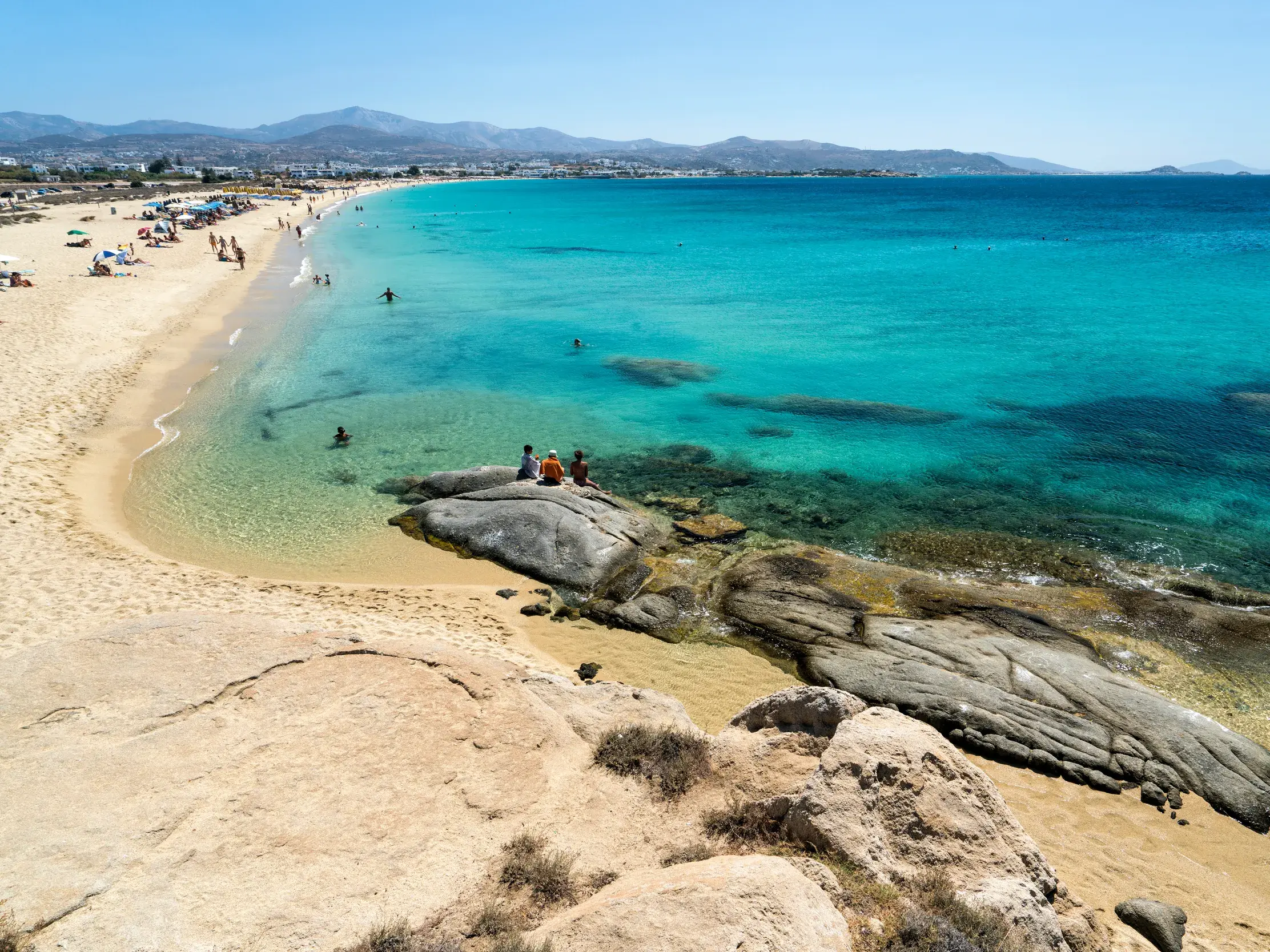 Weitläufiger Sandstrand auf Naxos mit kristallklarem, türkisfarbenem Wasser und Badegästen unter blauem Sommerhimmel.