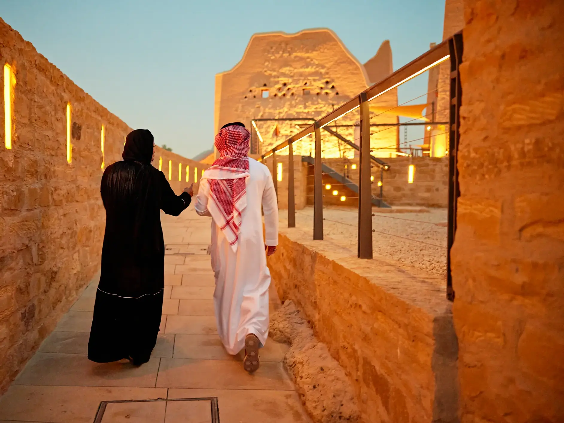 Paar in arabischer Tracht spaziert bei Sonnenuntergang durch eine Gasse der historischen Lehmstadt at-Turaif in Diriyah.