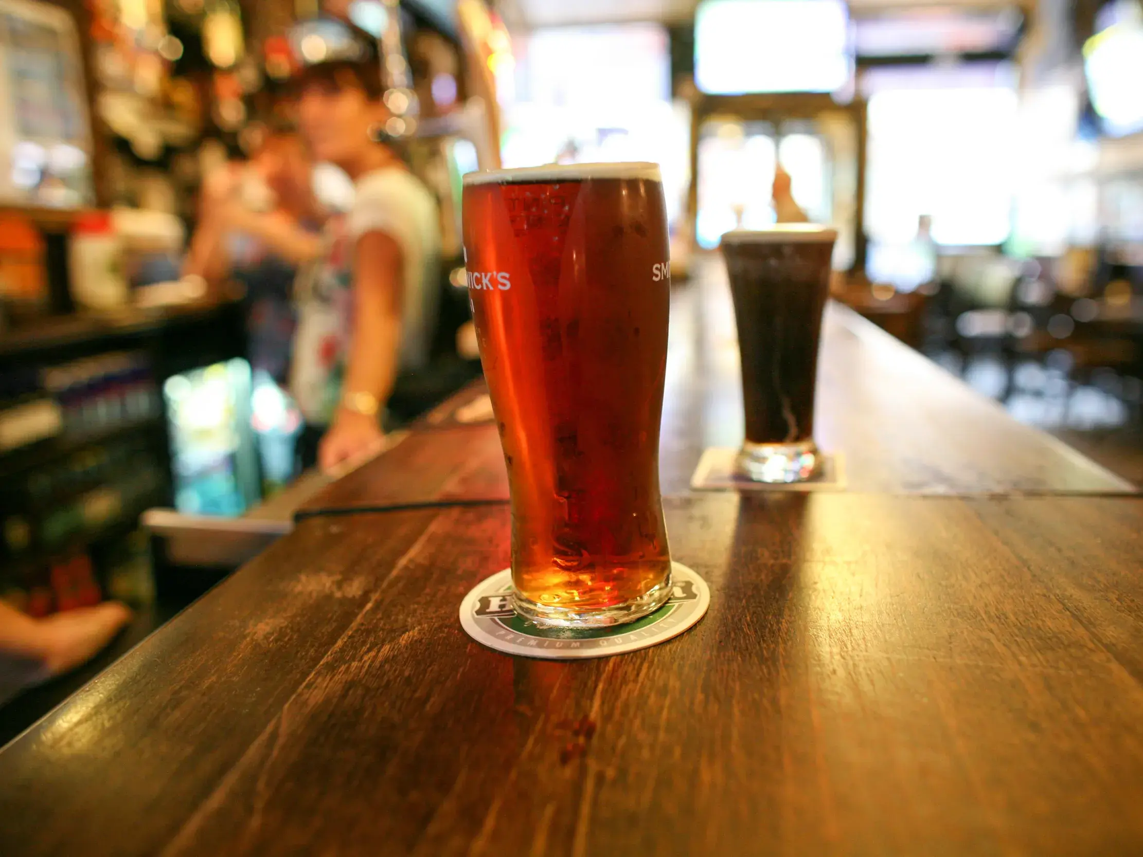 Zwei frisch gezapfte Biere, ein Ale und ein Stout, stehen auf dem Holztresen eines belebten irischen Pubs.