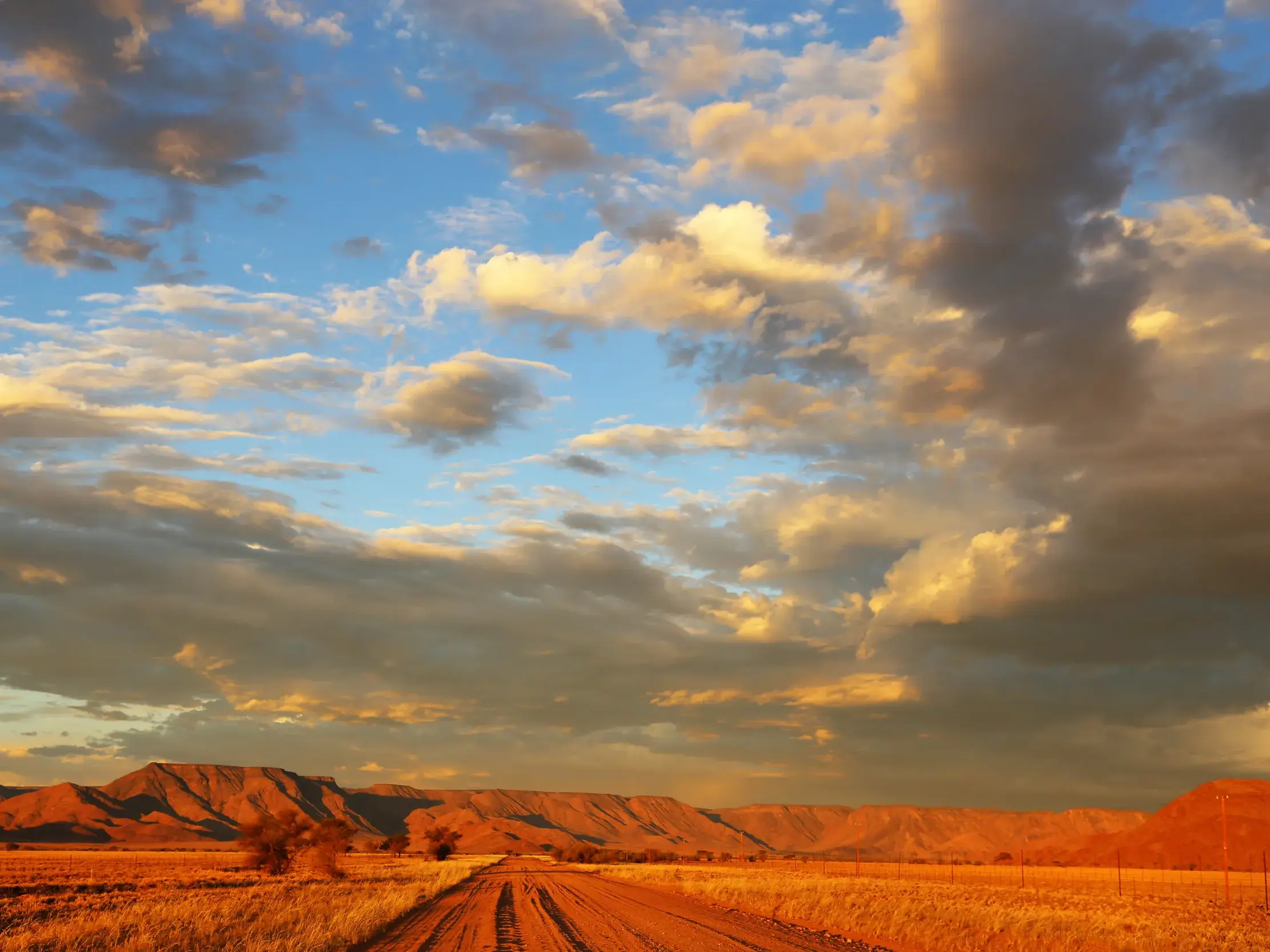 Eine rote Sandpiste führt durch die weite, goldene Wüstenlandschaft Namibias zu den Naukluftbergen bei Sonnenuntergang.