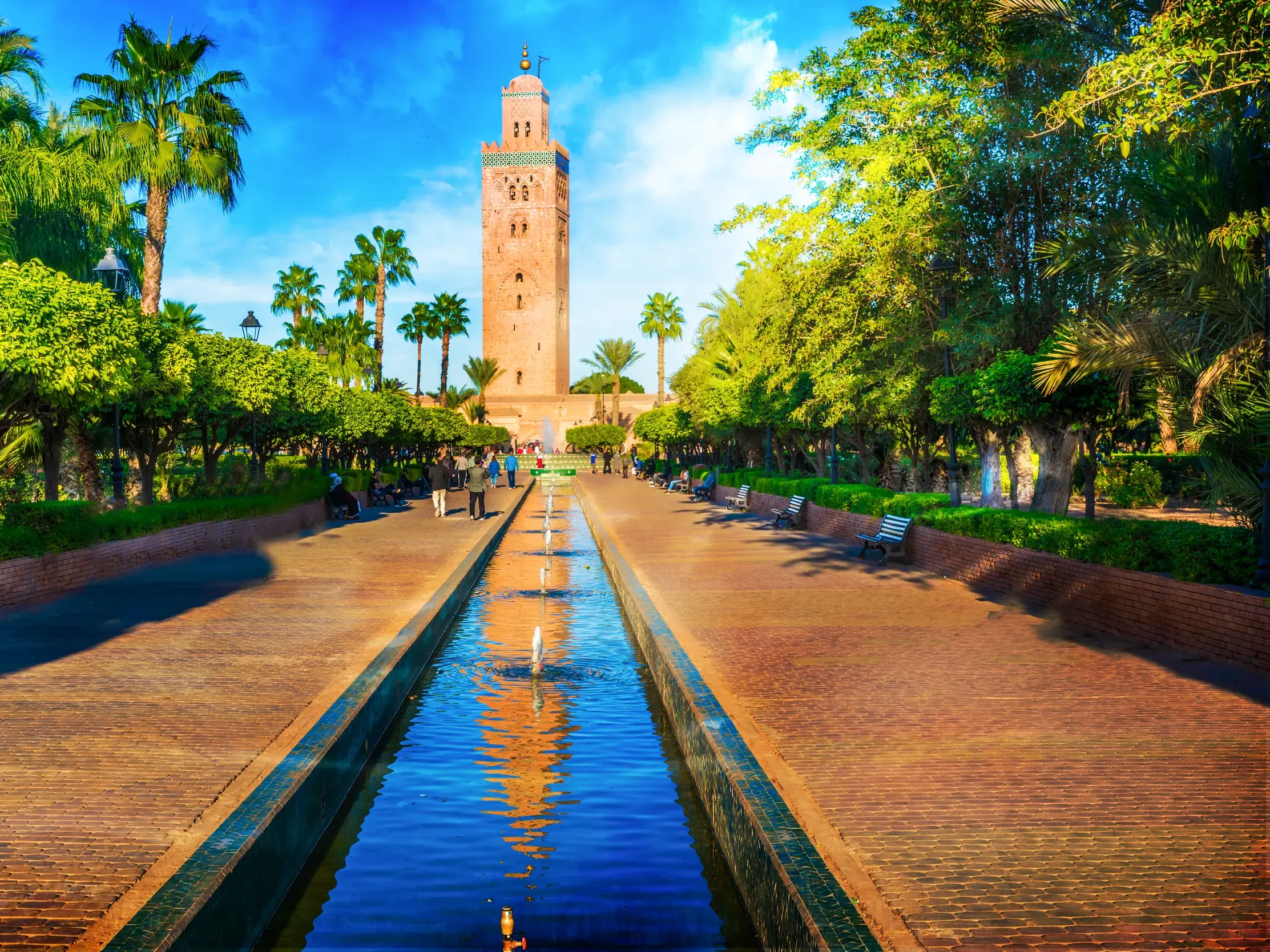 Langes Wasserbecken im Lalla Hasna Park führt zum Minarett der Koutoubia-Moschee in Marrakesch, umgeben von Palmen und Bäumen.