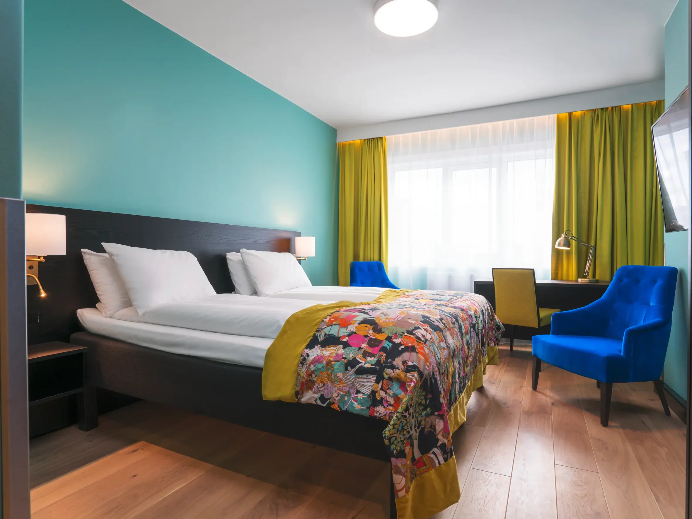 Stilvolles Doppelzimmer im Thon Hotel Europa in Oslo mit türkiser Wand, bunter Tagesdecke und blauem Samtsessel.