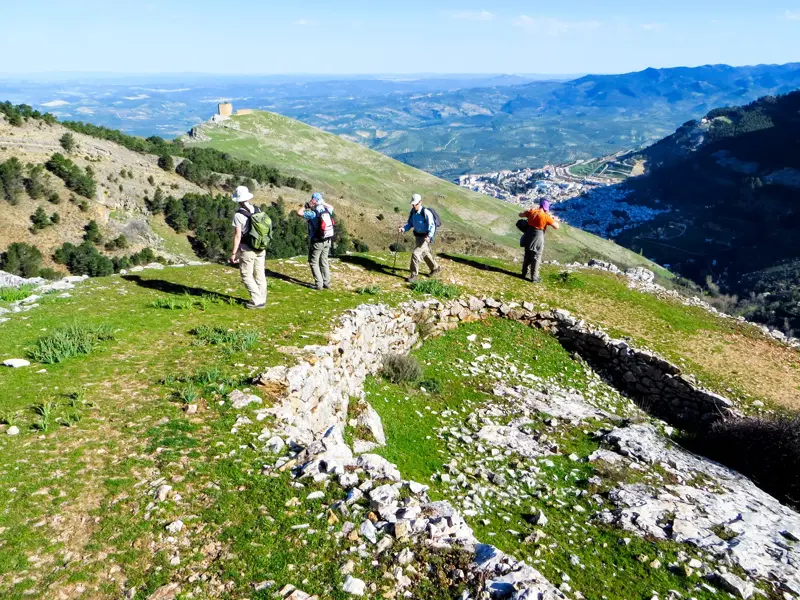 Vier Wanderer genießen auf einem grünen Berg in Andalusien die weite Aussicht auf ein weißes Dorf und eine Burgruine.
