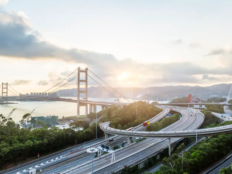 Eindrucksvolle Hängebrücke und verschlungene Autobahnen vor der Skyline einer chinesischen Metropole bei Sonnenaufgang.