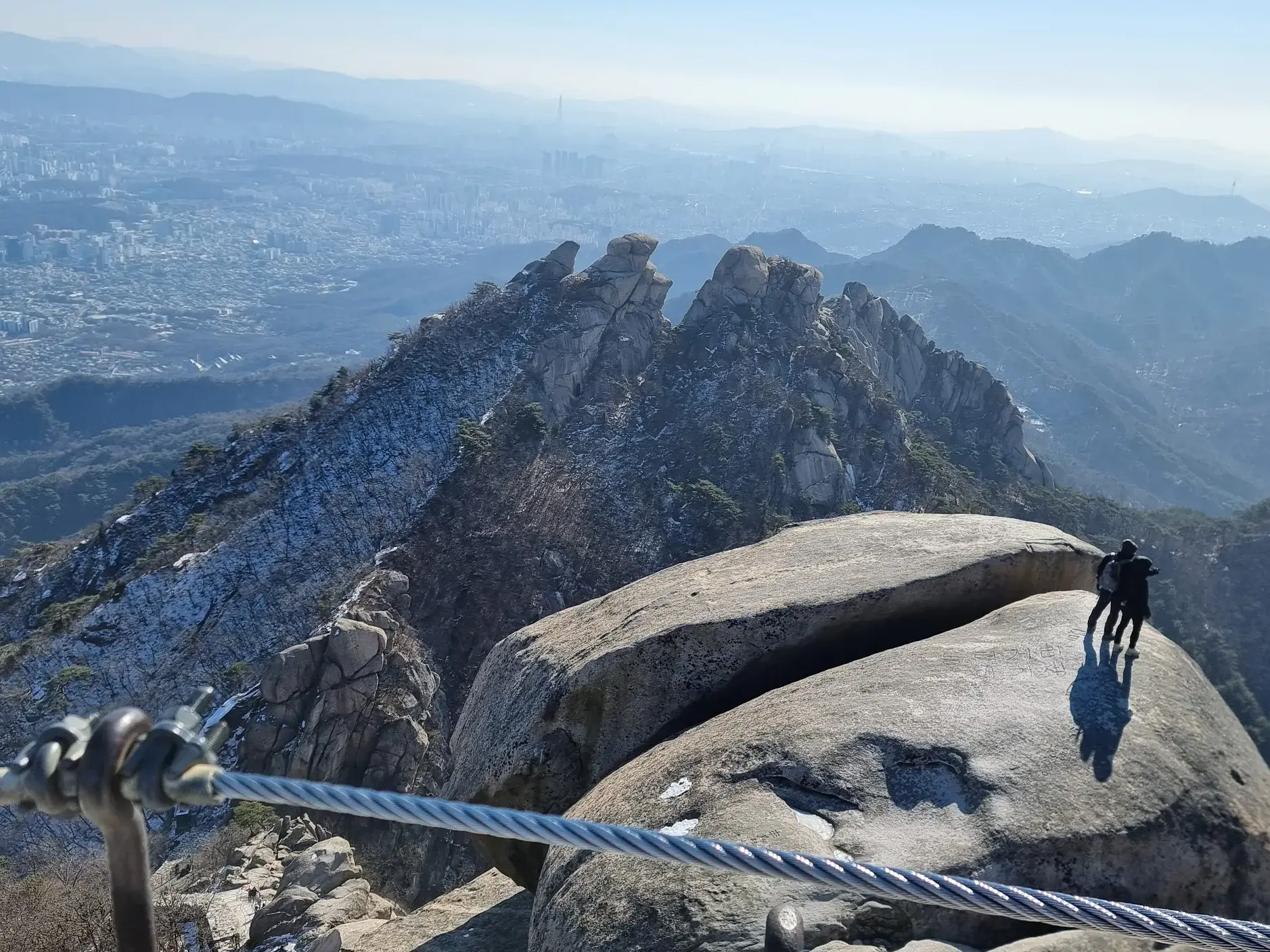 Zwei Wanderer auf dem felsigen Baegundae-Gipfel im Bukhansan-Nationalpark mit Panoramablick über die Metropole Seoul.