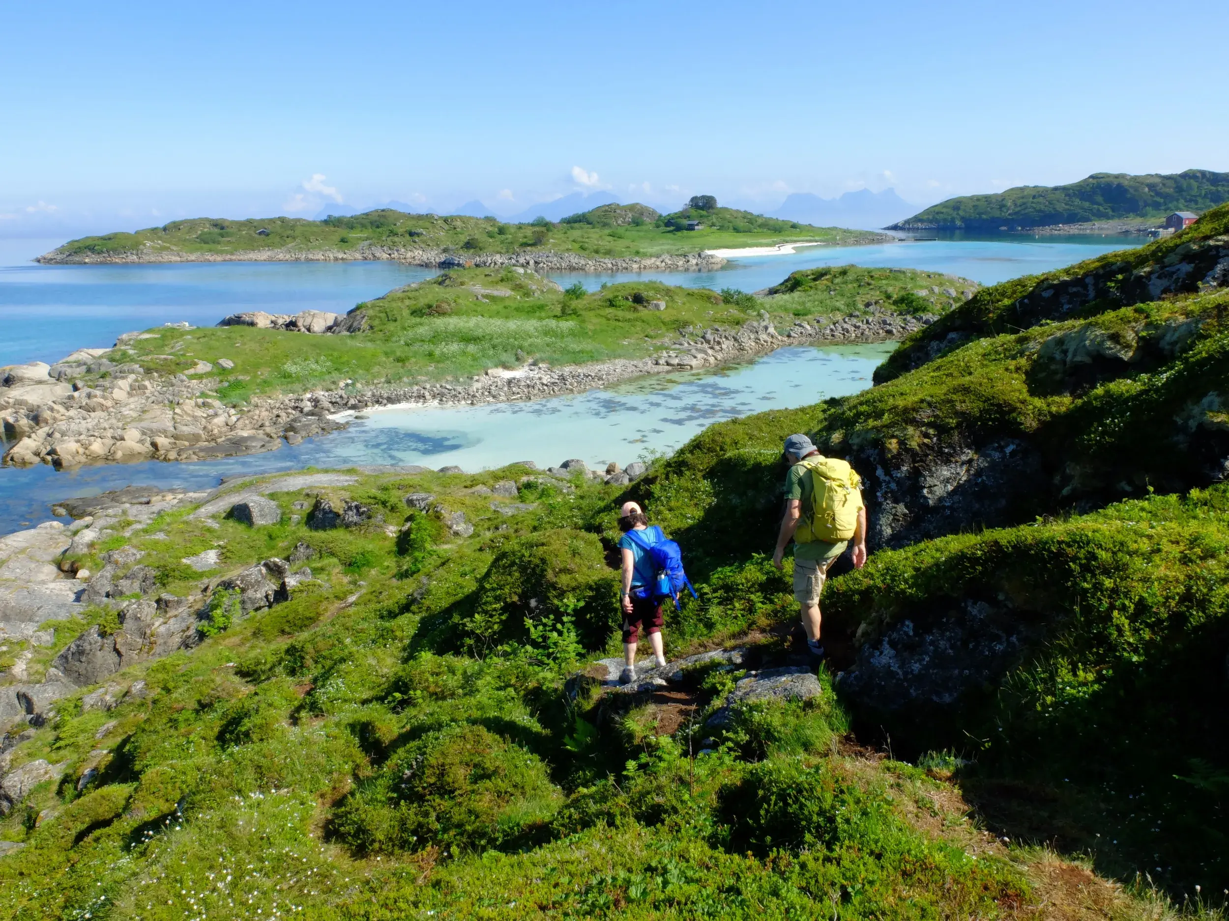 Zwei Wanderer auf einem Küstenpfad auf der Insel Skrova mit Blick auf türkisfarbene Buchten der Lofoten in Norwegen.