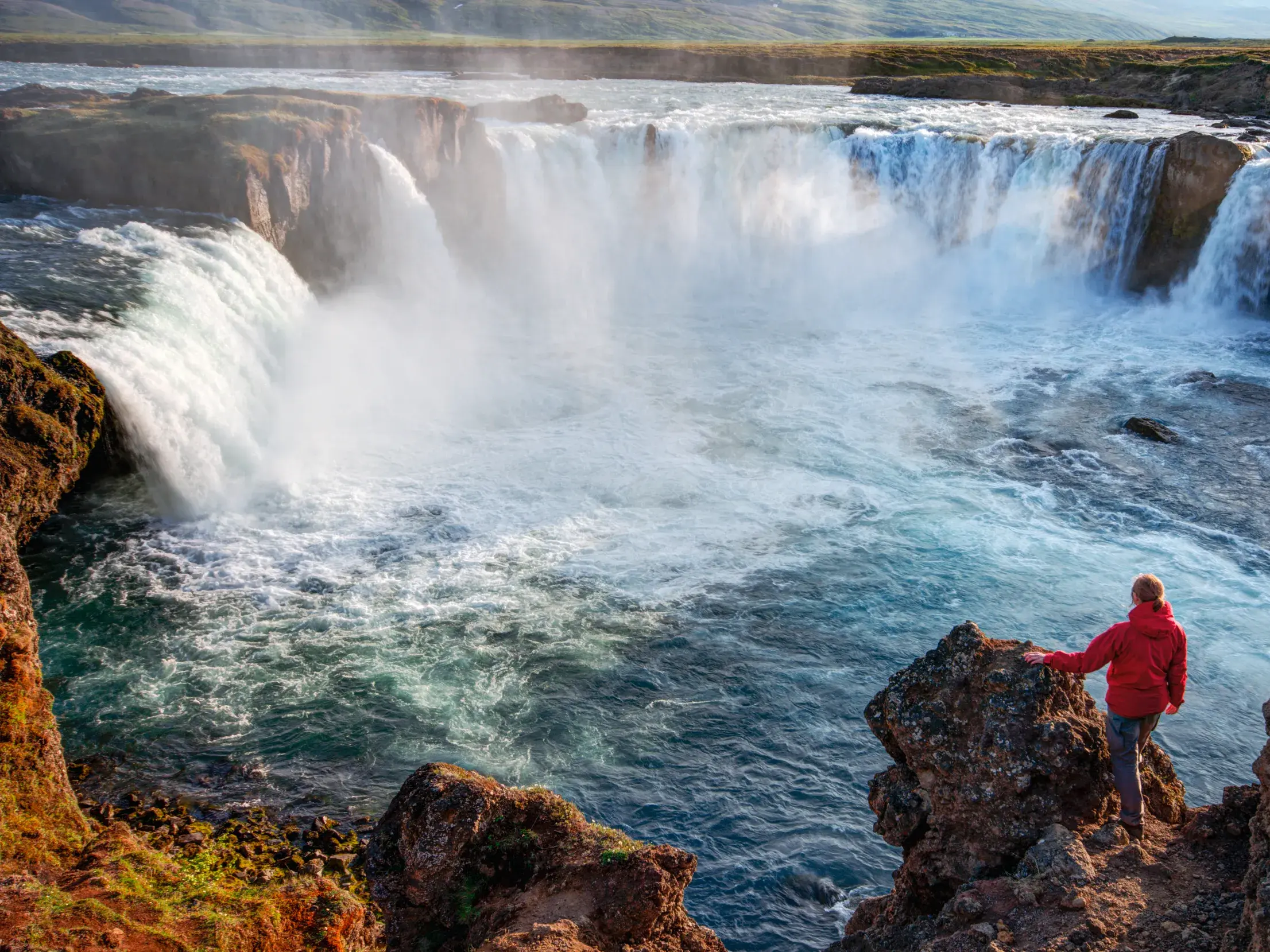 Person in roter Jacke blickt auf einer geführten Rundreise auf den tosenden Godafoss Wasserfall in Island.