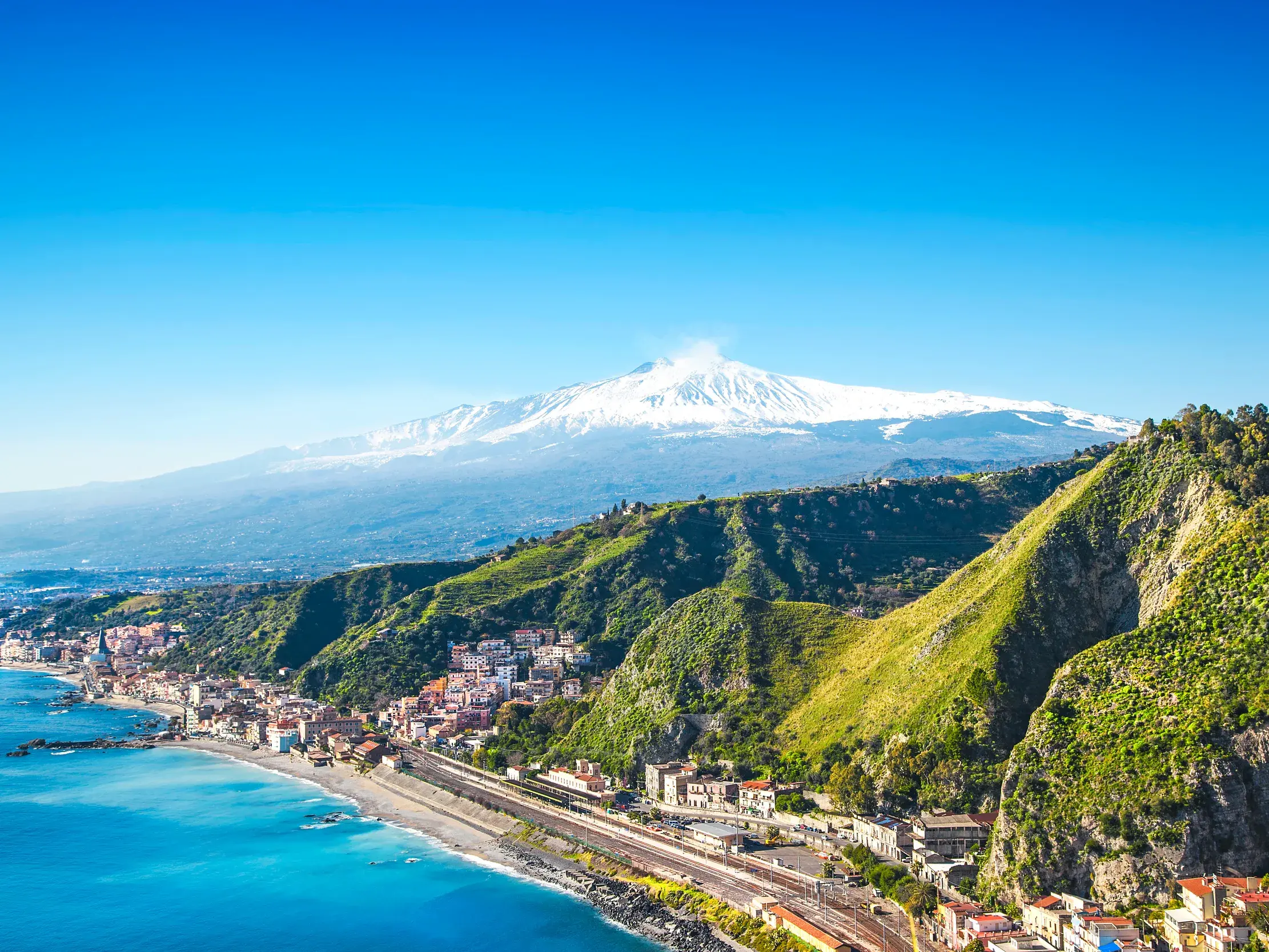 Spektakulärer Blick von Taormina auf die Küste, das Meer und den schneebedeckten, leicht rauchenden Vulkan Ätna in Sizilien.