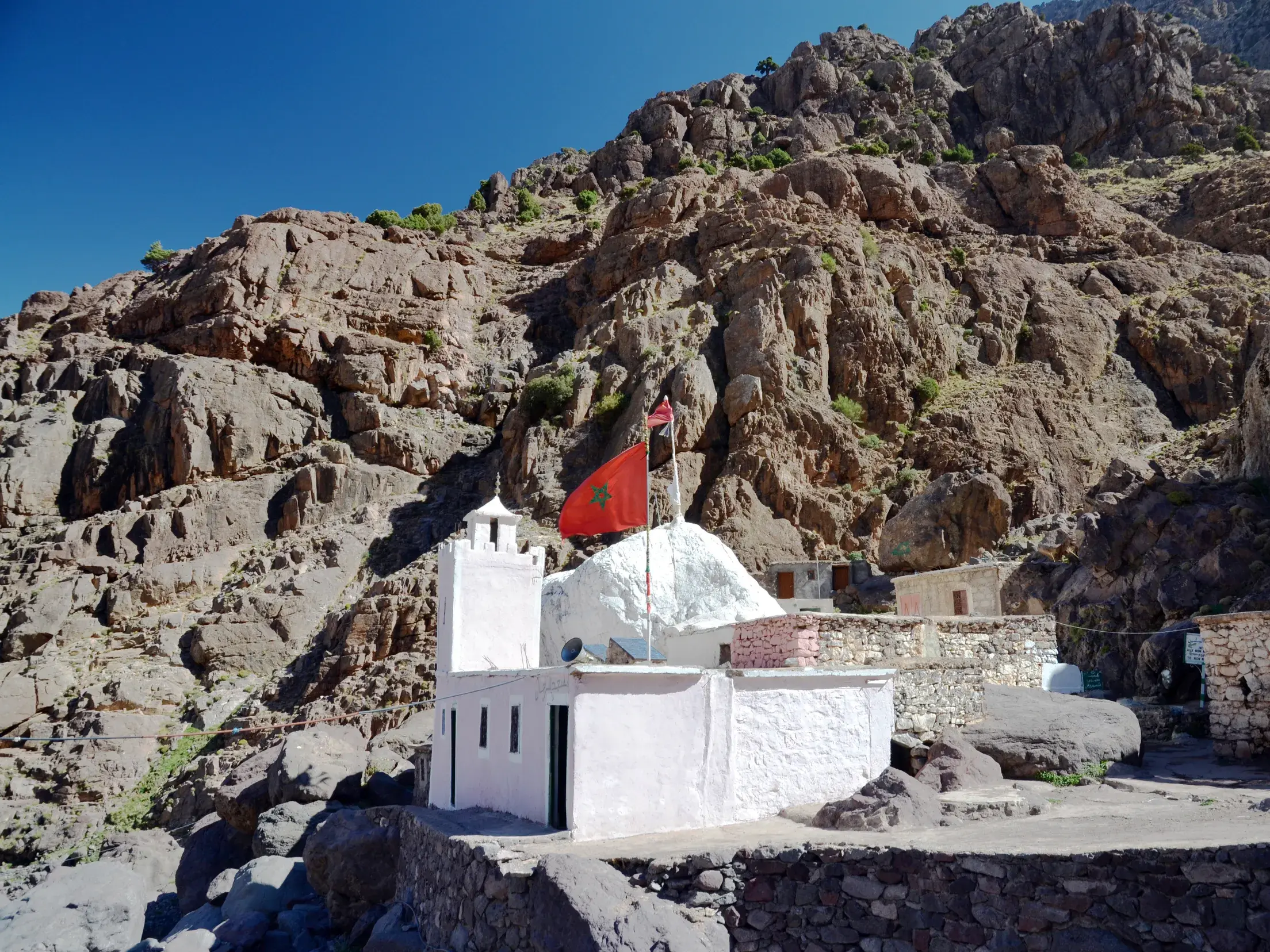 Der weiße Pilgerschrein Sidi Chamharouch mit Marokko-Flagge auf dem Trekkingpfad zum Jebel Toubkal im Hohen Atlas.