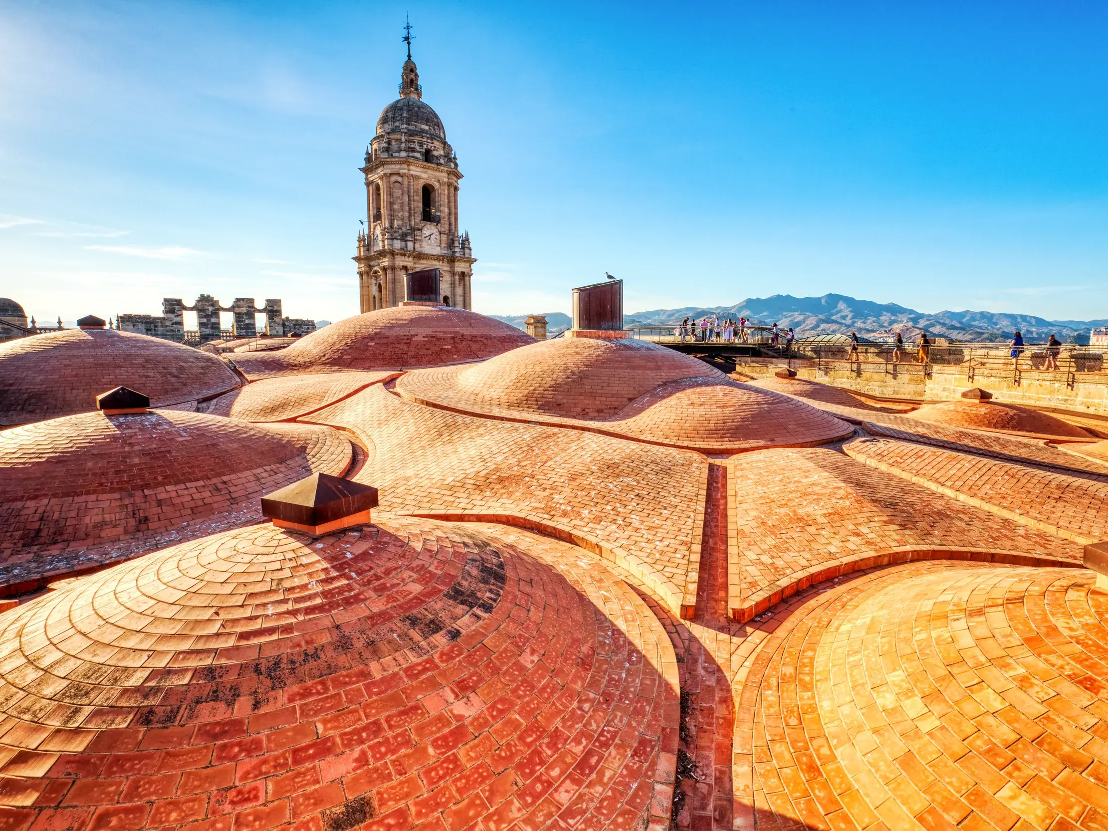 Blick von den gewölbten Terrakotta-Dächern der Kathedrale in Málaga auf den Glockenturm und die Berglandschaft Andalusiens.