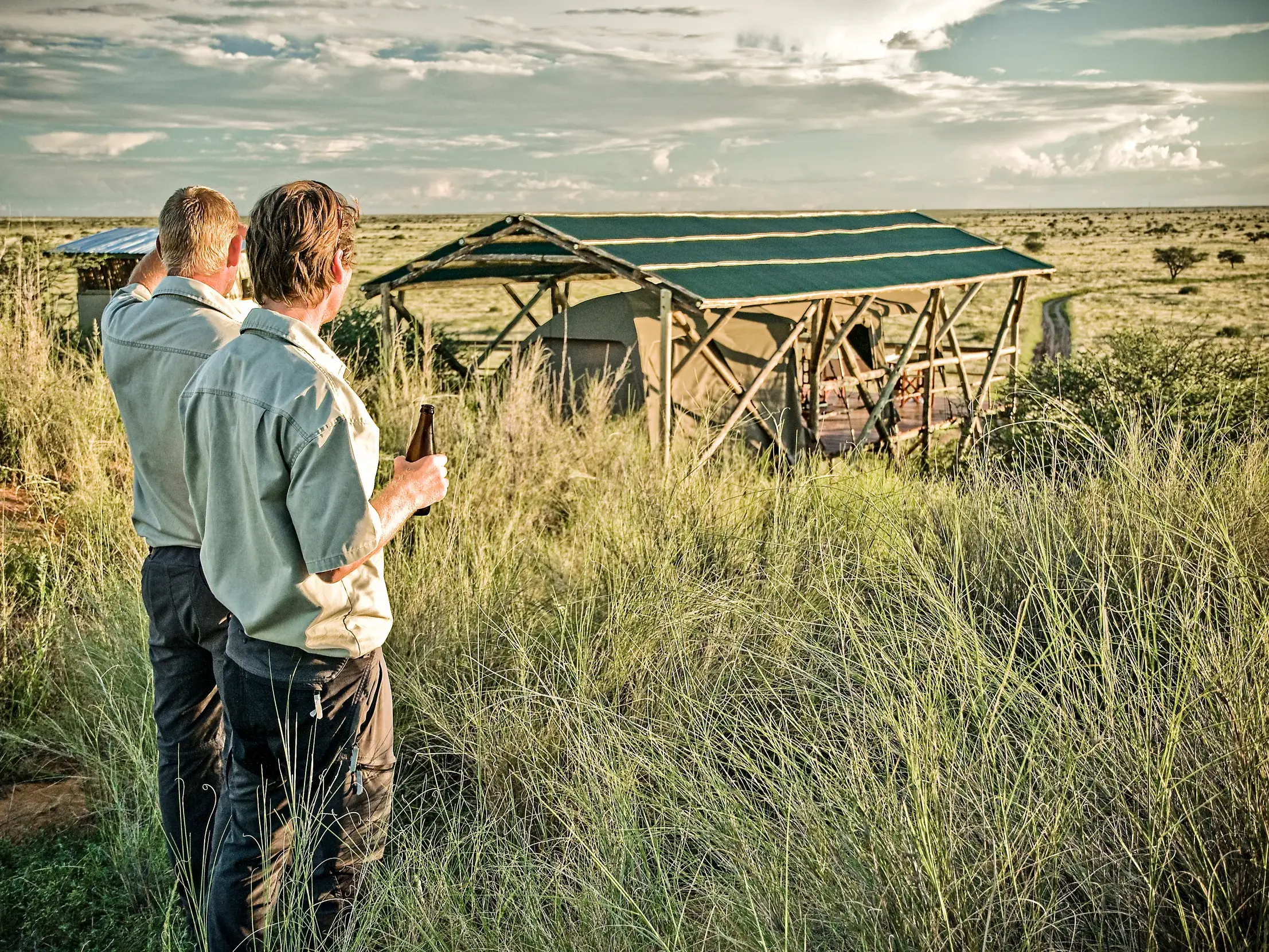 Zwei Männer genießen mit einem Bier den Sundowner vor einem Zeltcamp in der weiten Savanne der Kalahari in Namibia.