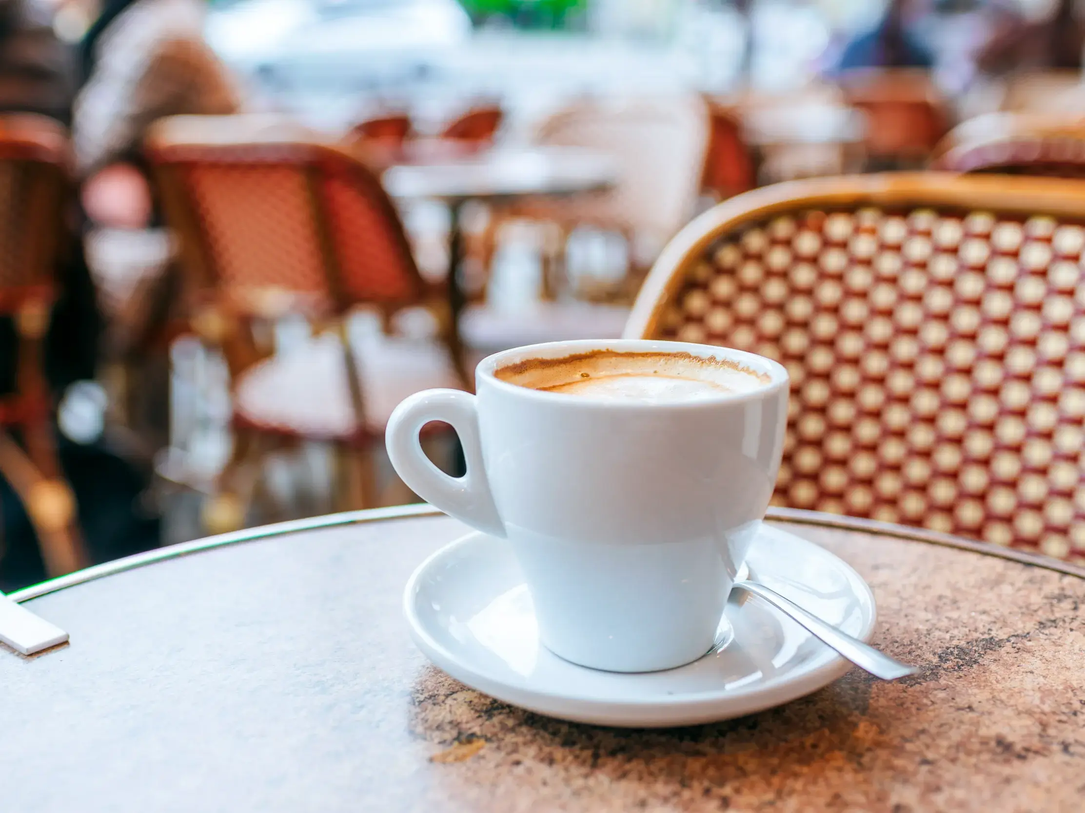 Nahaufnahme einer weißen Kaffeetasse auf einem Marmortisch in einem typischen Straßencafé in Paris mit Korbstühlen im Hintergrund.