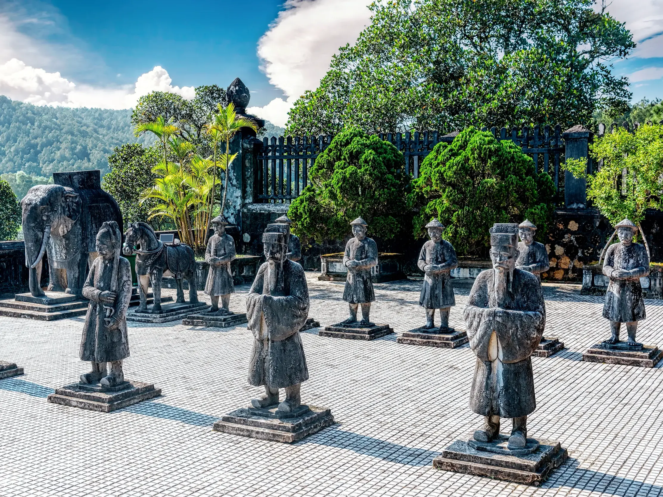 Die steinerne Ehrengarde mit Soldaten und Elefanten bewacht das Kaisergrab von Minh Mang bei Hue vor einer grünen Hügellandschaft.