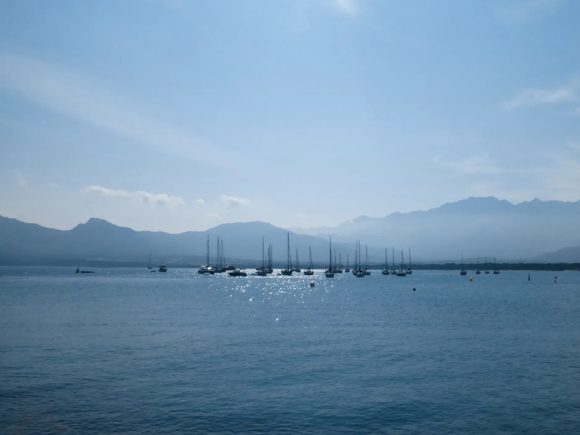 Panoramablick über die sonnenglitzernde Bucht von Calvi, in der Segelboote vor der Bergkulisse Korsikas ankern.