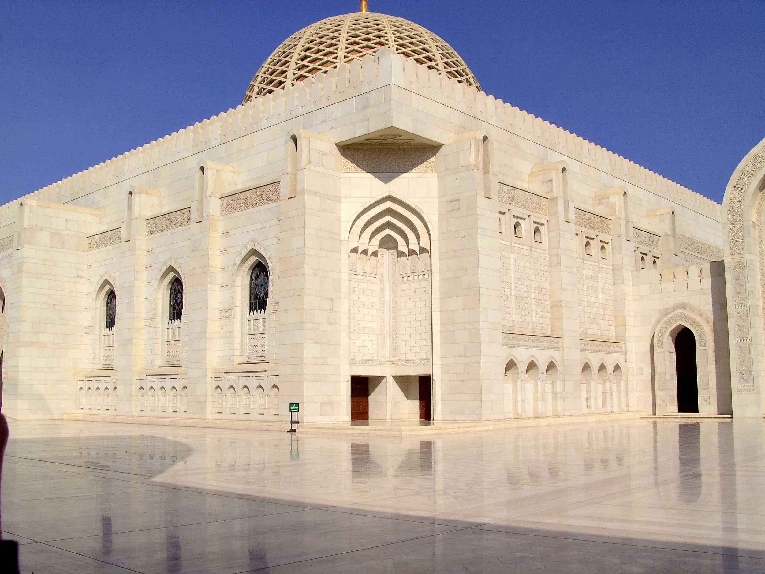 Fassade der prunkvollen Sultan-Qabus-Moschee in Maskat, deren weißer Marmor sich auf dem polierten Vorplatz spiegelt.