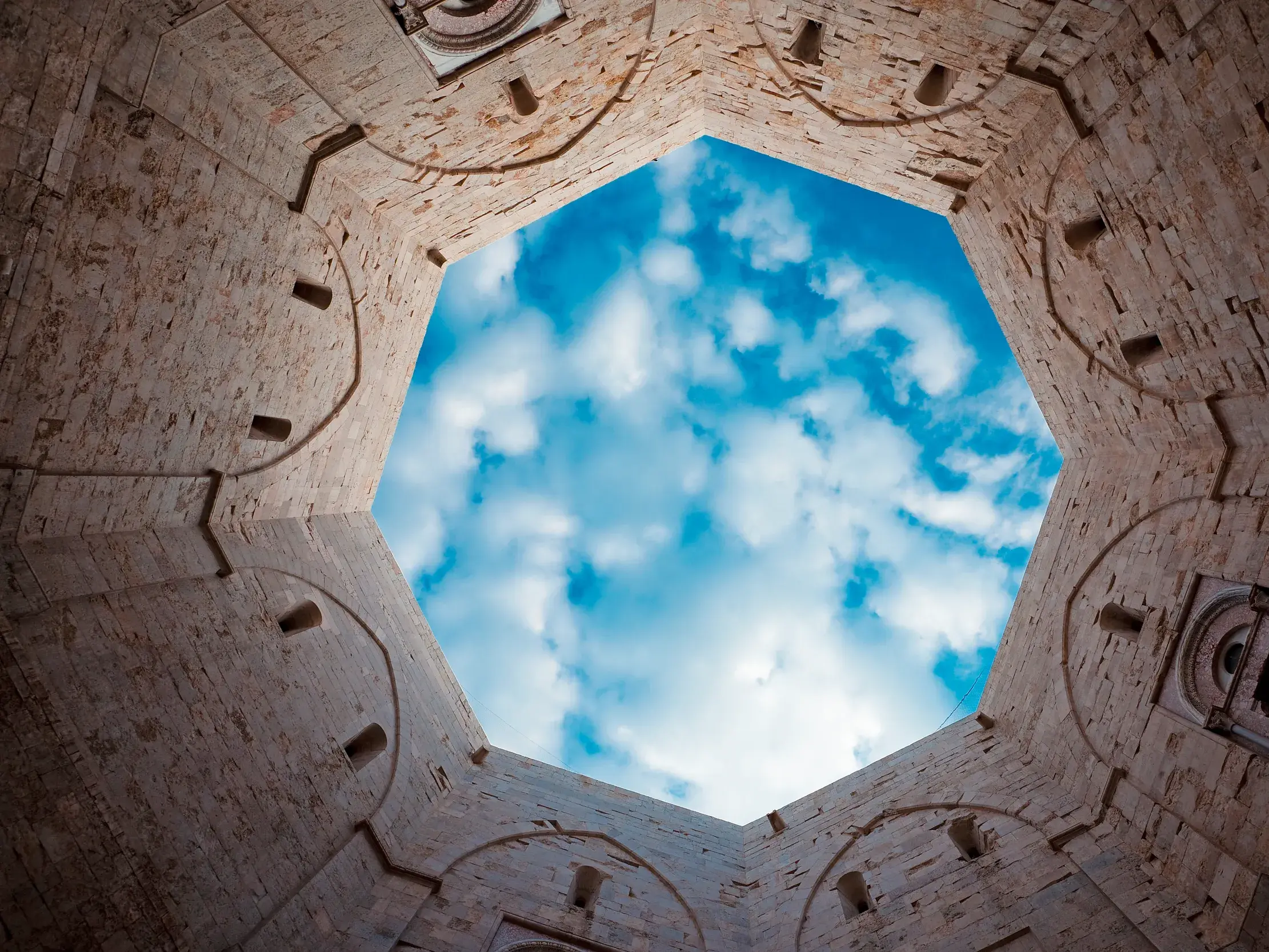 Blick aus dem achteckigen Innenhof des Castel del Monte in Apulien durch die Deckenöffnung zum blauen, bewölkten Himmel.