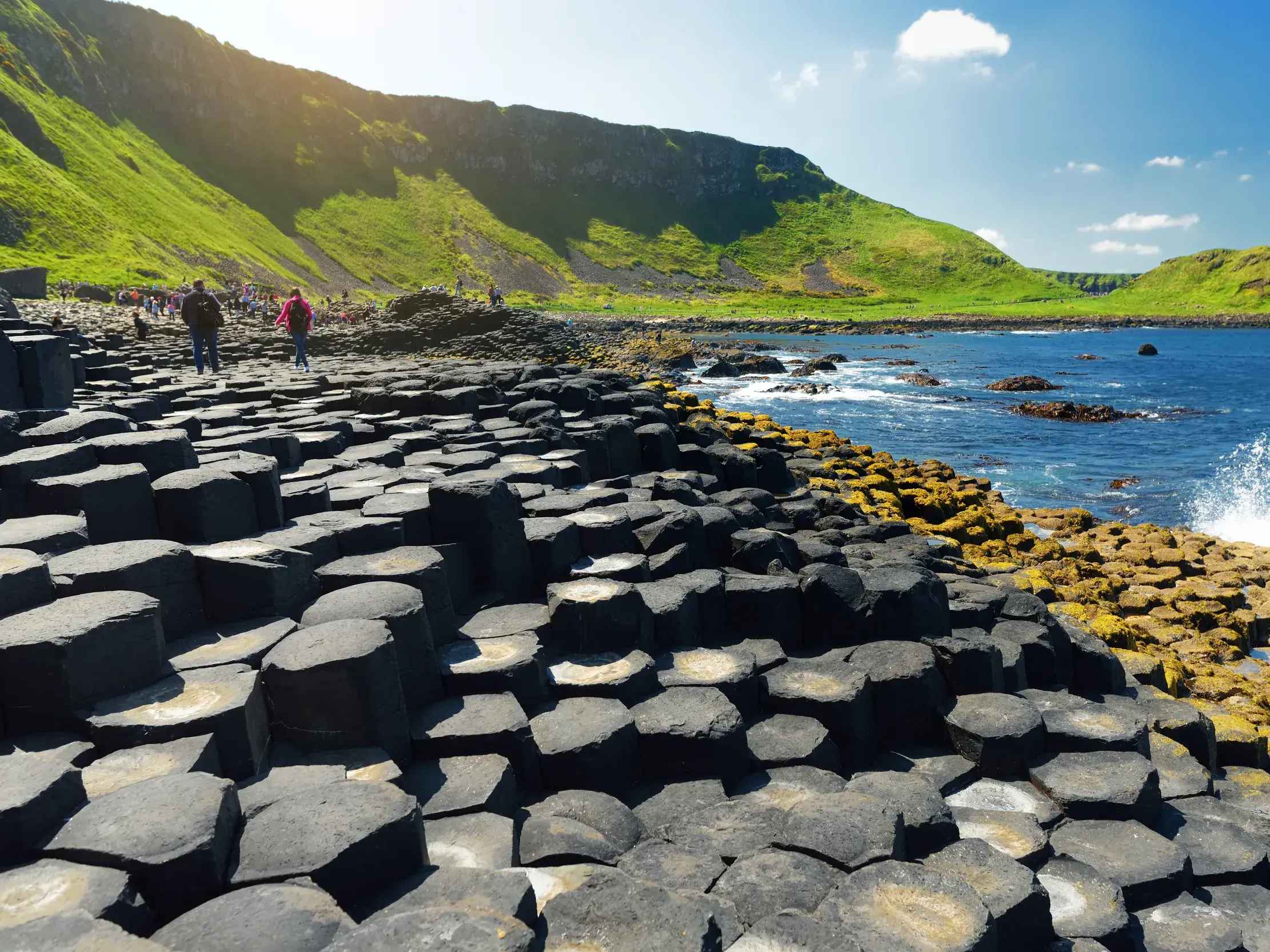 Wanderer auf den markanten Basaltsäulen des Giant's Causeway an der sonnigen Küste Nordirlands vor grünen Klippen.