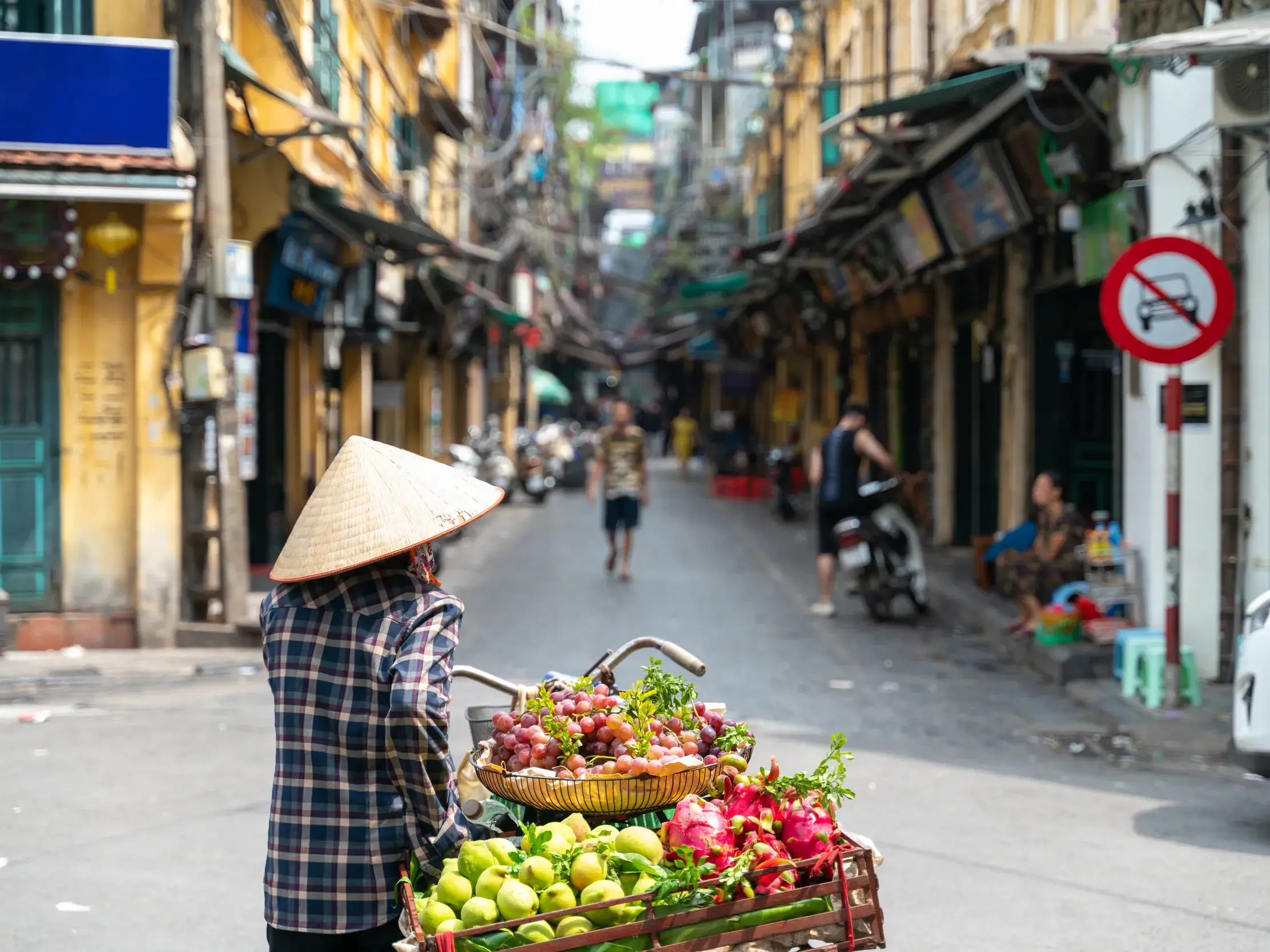 Vietnamesische Obstverkäuferin mit Kegelhut schiebt ihr Fahrrad mit frischen Früchten durch eine Gasse im Alten Viertel von Hanoi.