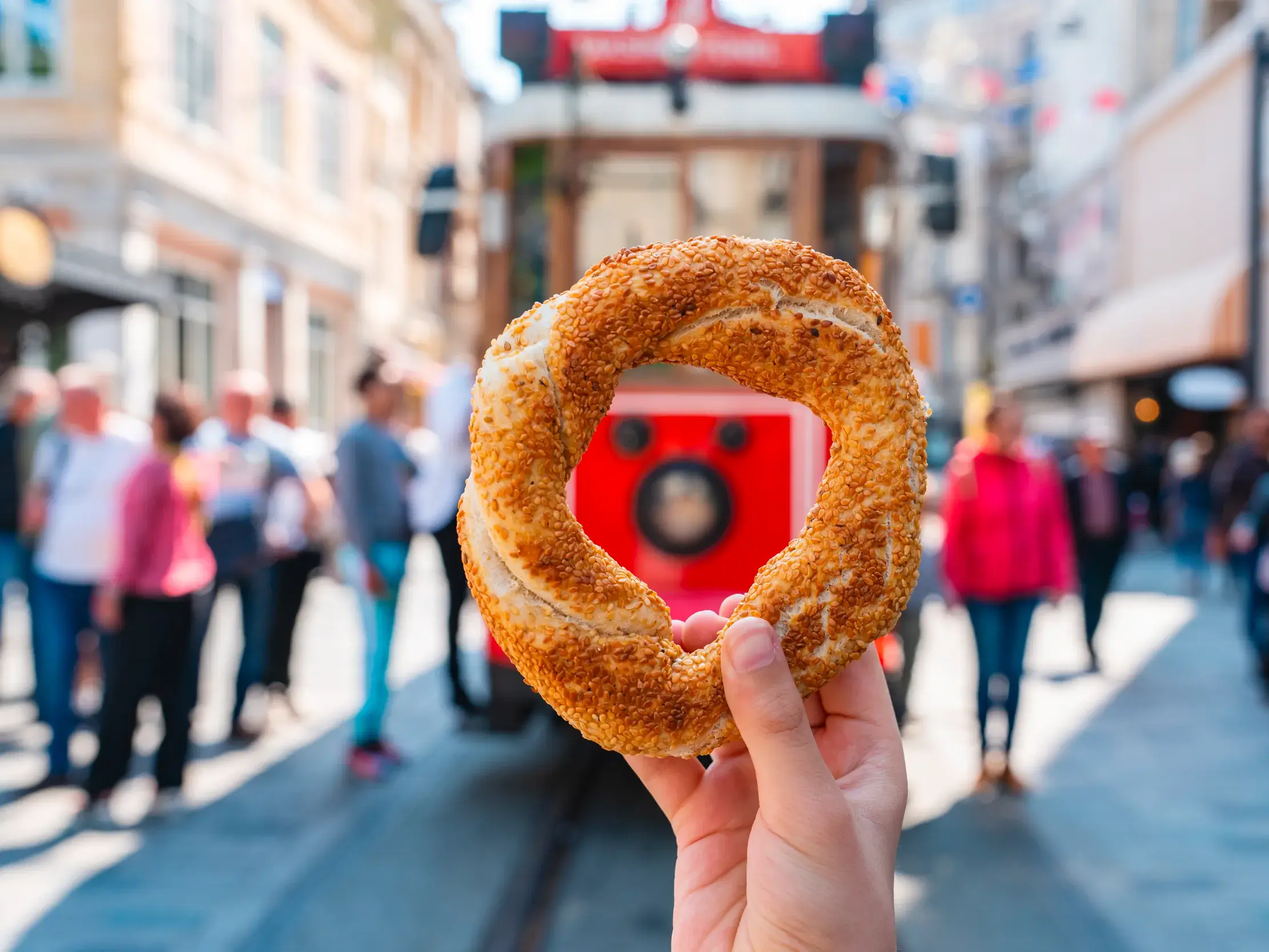 Ein Sesamring, Simit genannt, wird in die Kamera gehalten, im Hintergrund die belebte Einkaufsstraße Istiklal Caddesi in Istanbul.