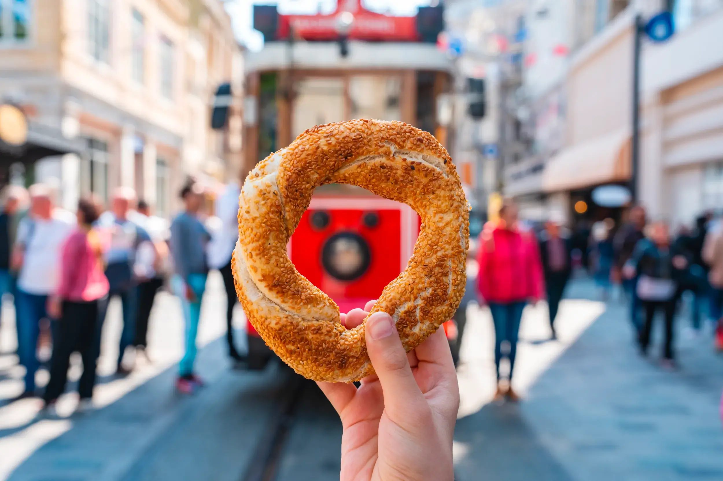Ein Sesamring, Simit genannt, wird in die Kamera gehalten, im Hintergrund die belebte Einkaufsstraße Istiklal Caddesi in Istanbul.