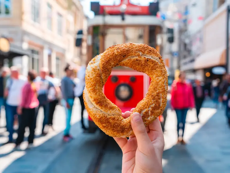 Ein Sesamring, Simit genannt, wird in die Kamera gehalten, im Hintergrund die belebte Einkaufsstraße Istiklal Caddesi in Istanbul.