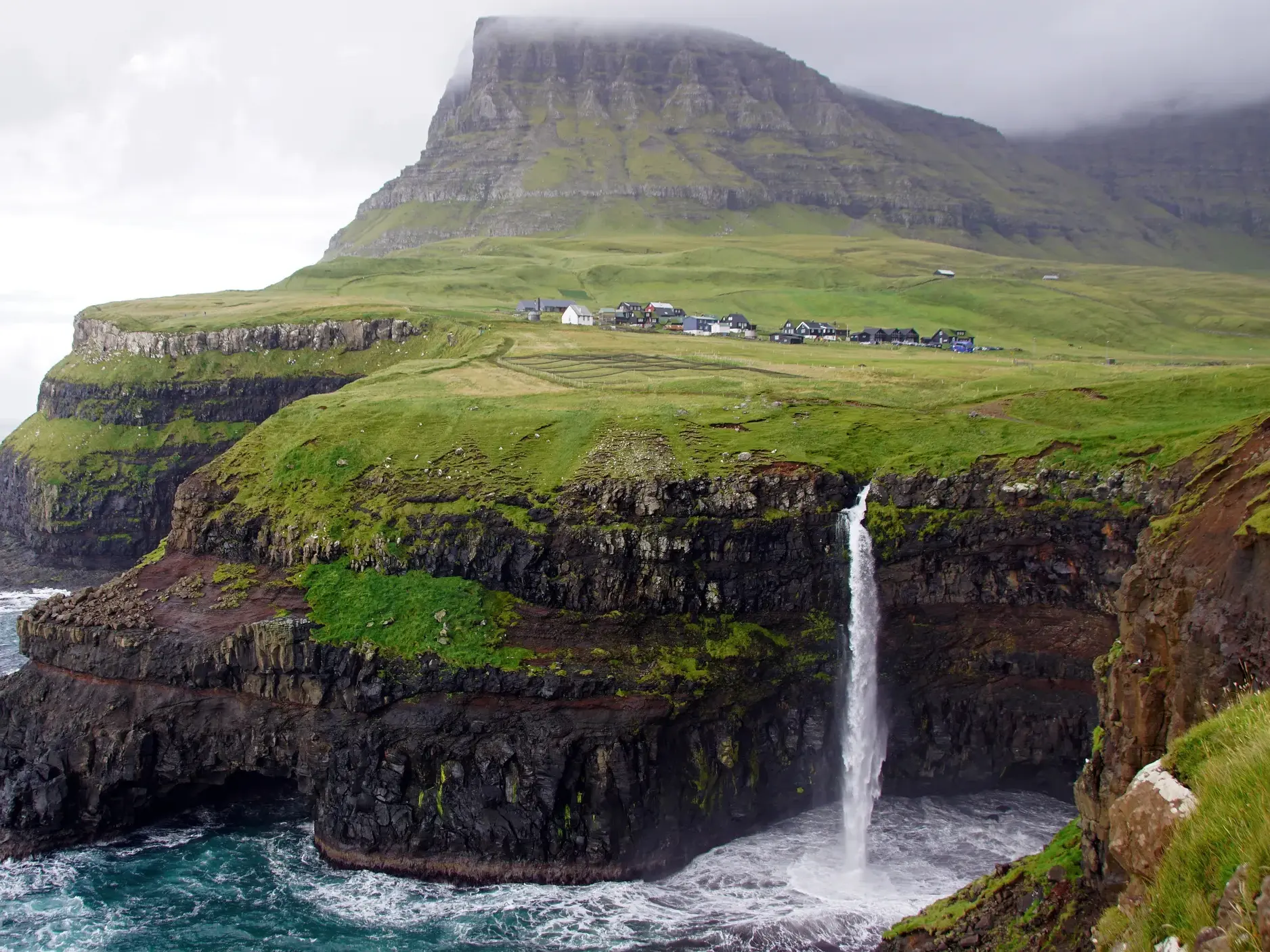 Der Múlafossur Wasserfall stürzt auf der Insel Vágar von einer grünen Steilklippe ins Meer, im Hintergrund das Dorf Gásadalur.