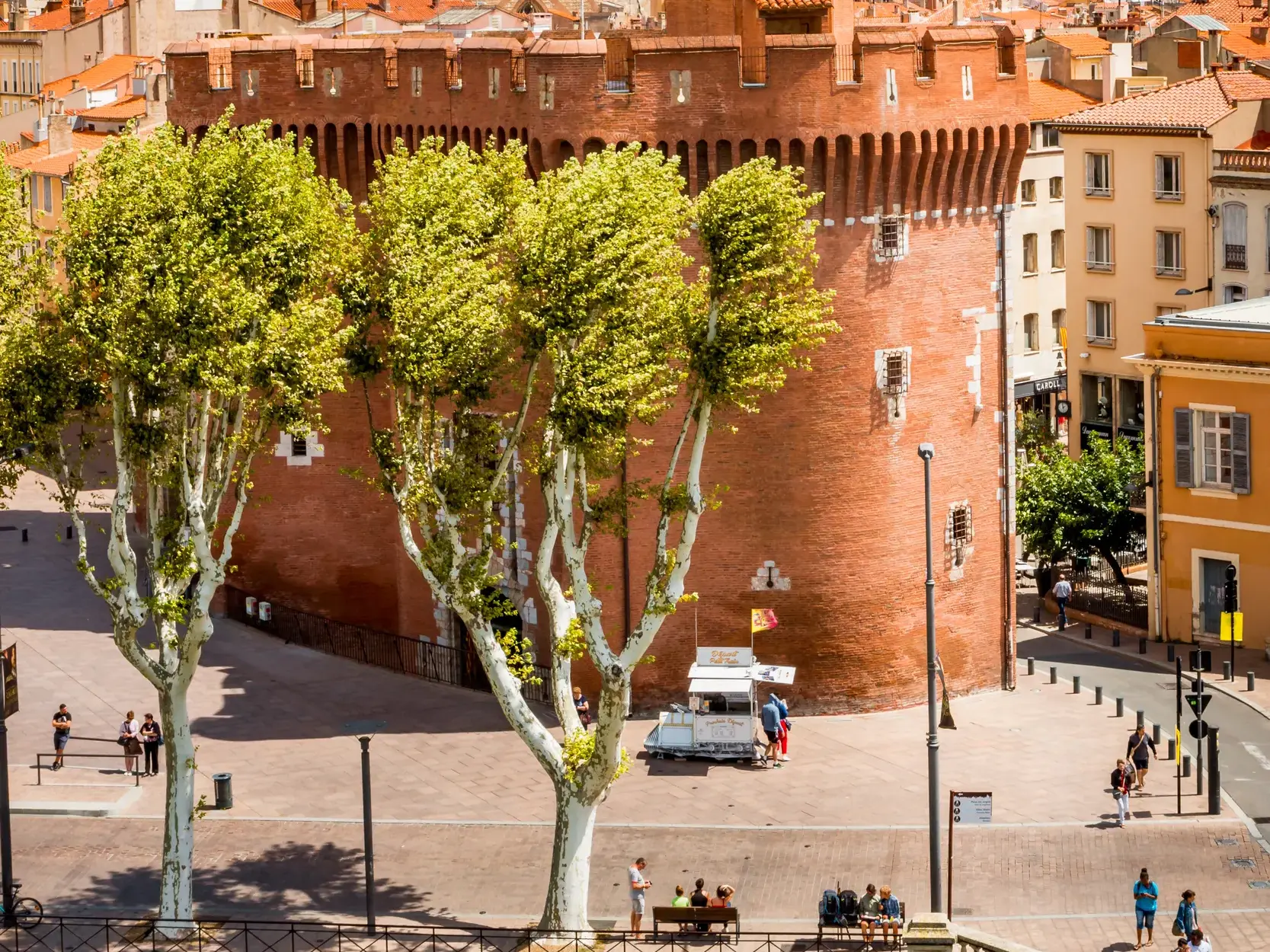 Sonniger Platz vor dem historischen Stadttor Castillet in Perpignan mit hohen Platanen und flanierenden Passanten.
