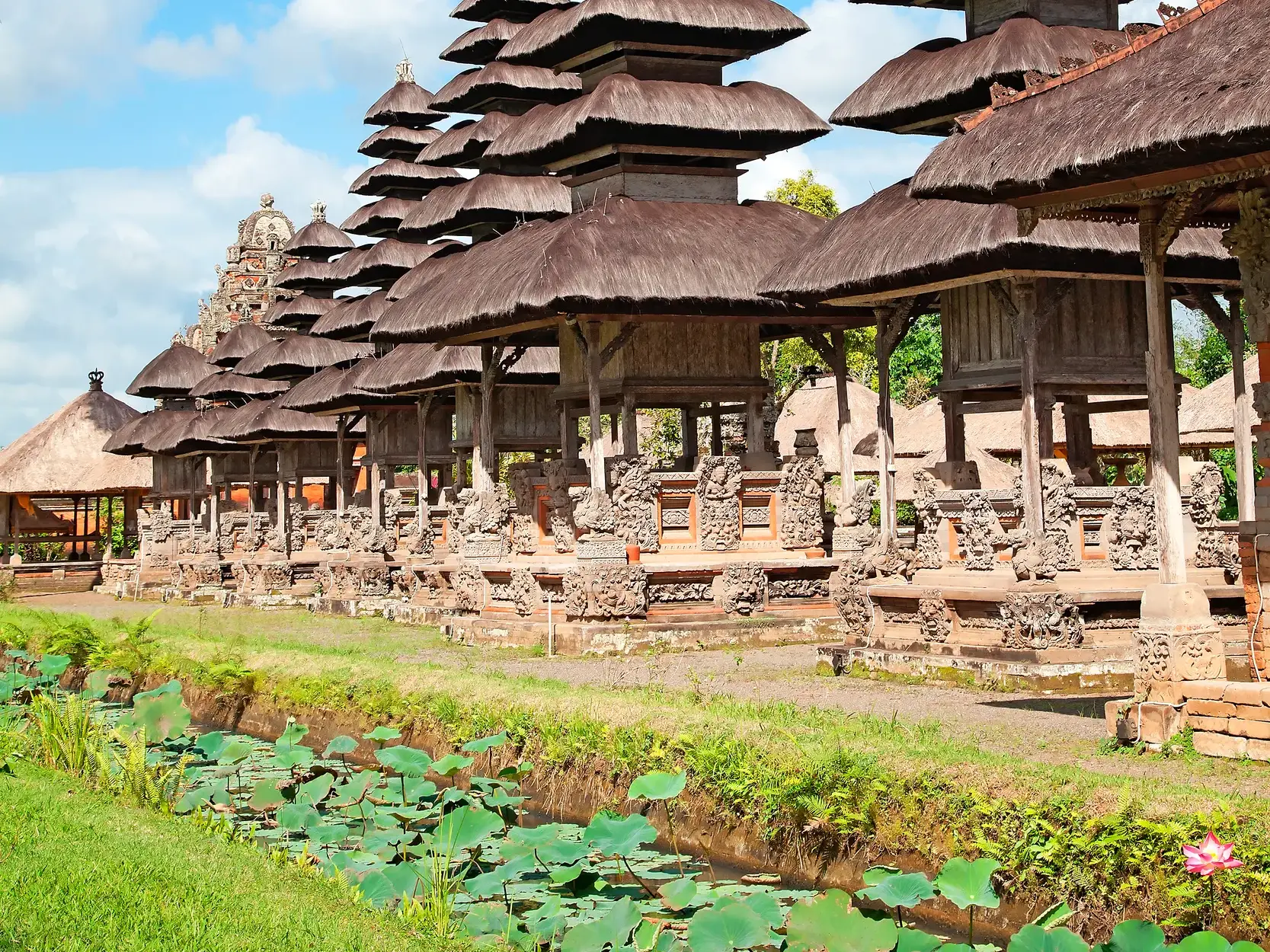 Die gestuften Meru-Türme des Taman-Ayun-Tempels auf Bali ragen an einem sonnigen Tag über einen Wassergraben mit Lotusblättern.
