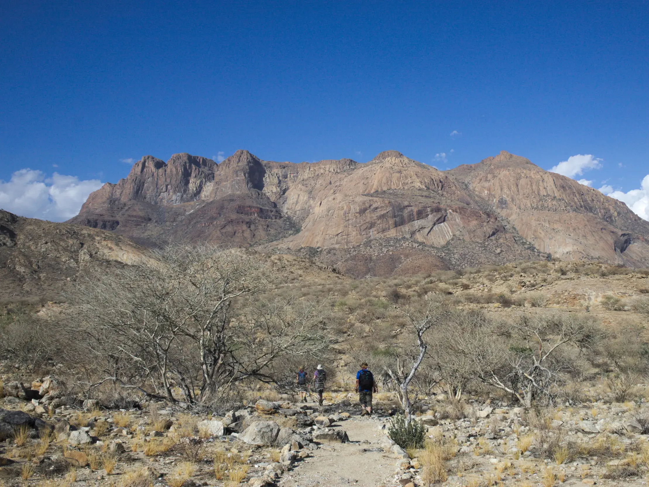 Eine kleine Wandergruppe erkundet zu Fuß die trockene Felslandschaft vor dem massiven Erongo-Gebirge in Namibia.