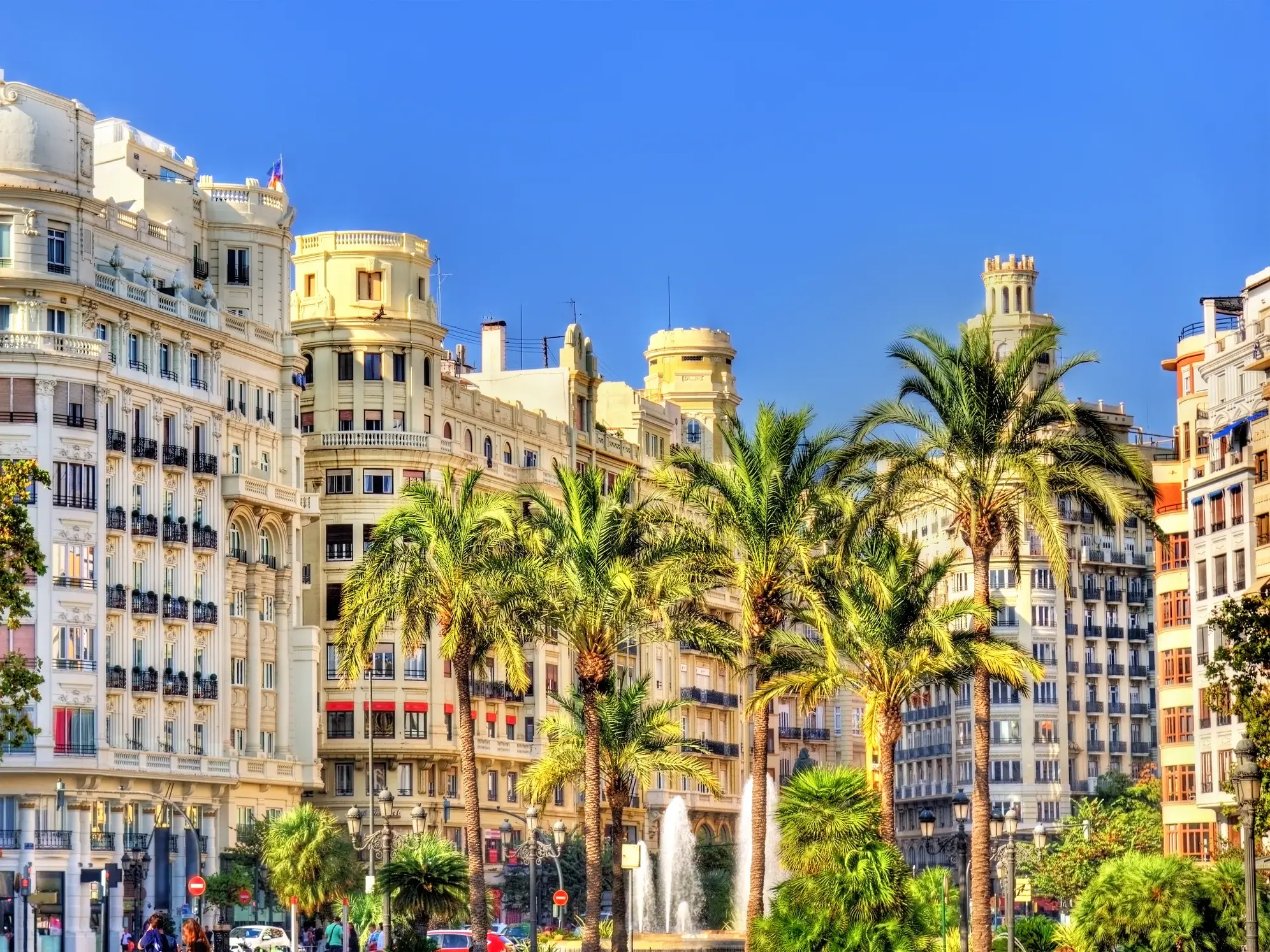 Historische Prachtbauten in der Altstadt von Valencia, gesäumt von Palmen und einem Brunnen unter strahlend blauem Himmel.