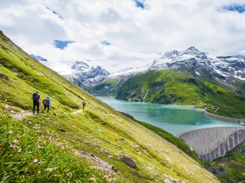 Eine Wandergruppe auf einem Pfad oberhalb des türkisblauen Mooserboden-Stausees im Nationalpark Hohe Tauern.