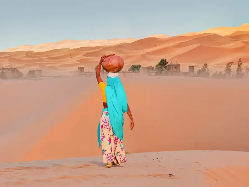 Frau mit Tonkrug auf dem Kopf wandert durch die weite Wüstenlandschaft der Sanddünen des Erg Chebbi.