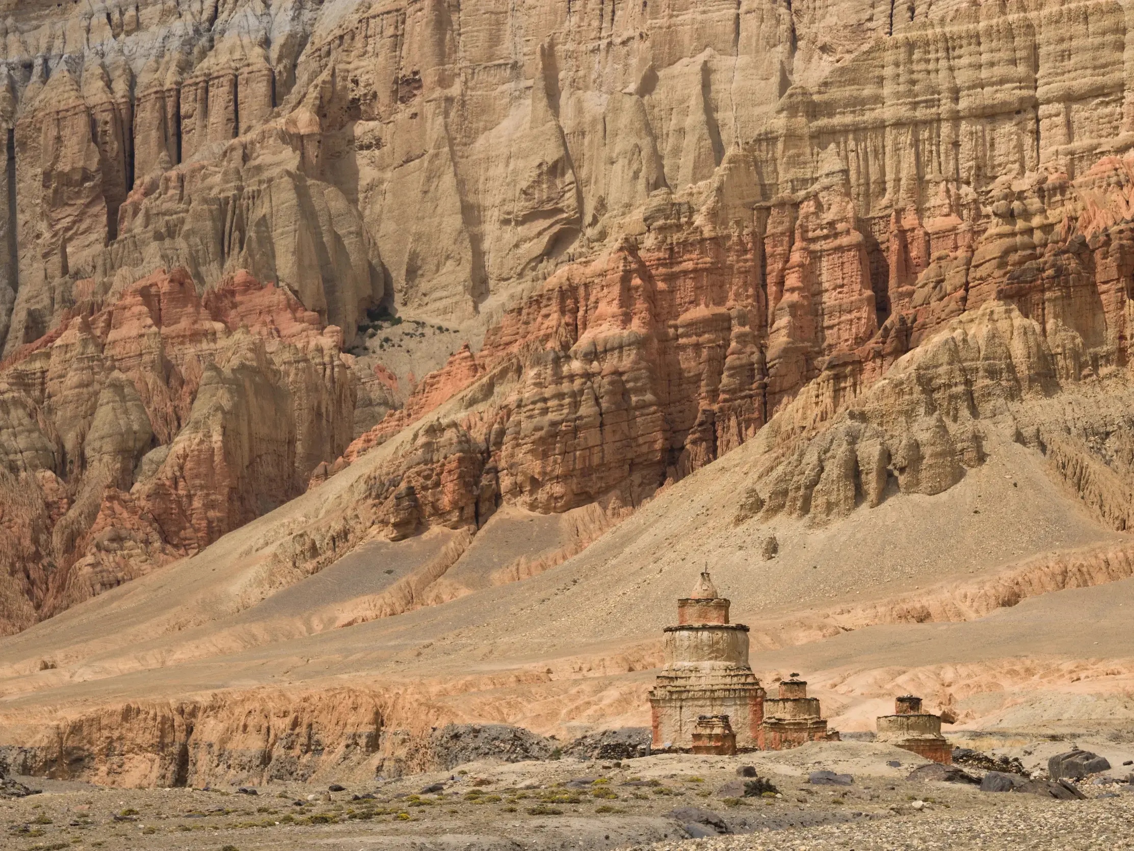 Steinerne Chörten vor einer imposanten, rot-braunen Felswand in der wüstenähnlichen Landschaft des Oberen Mustang in Nepal.