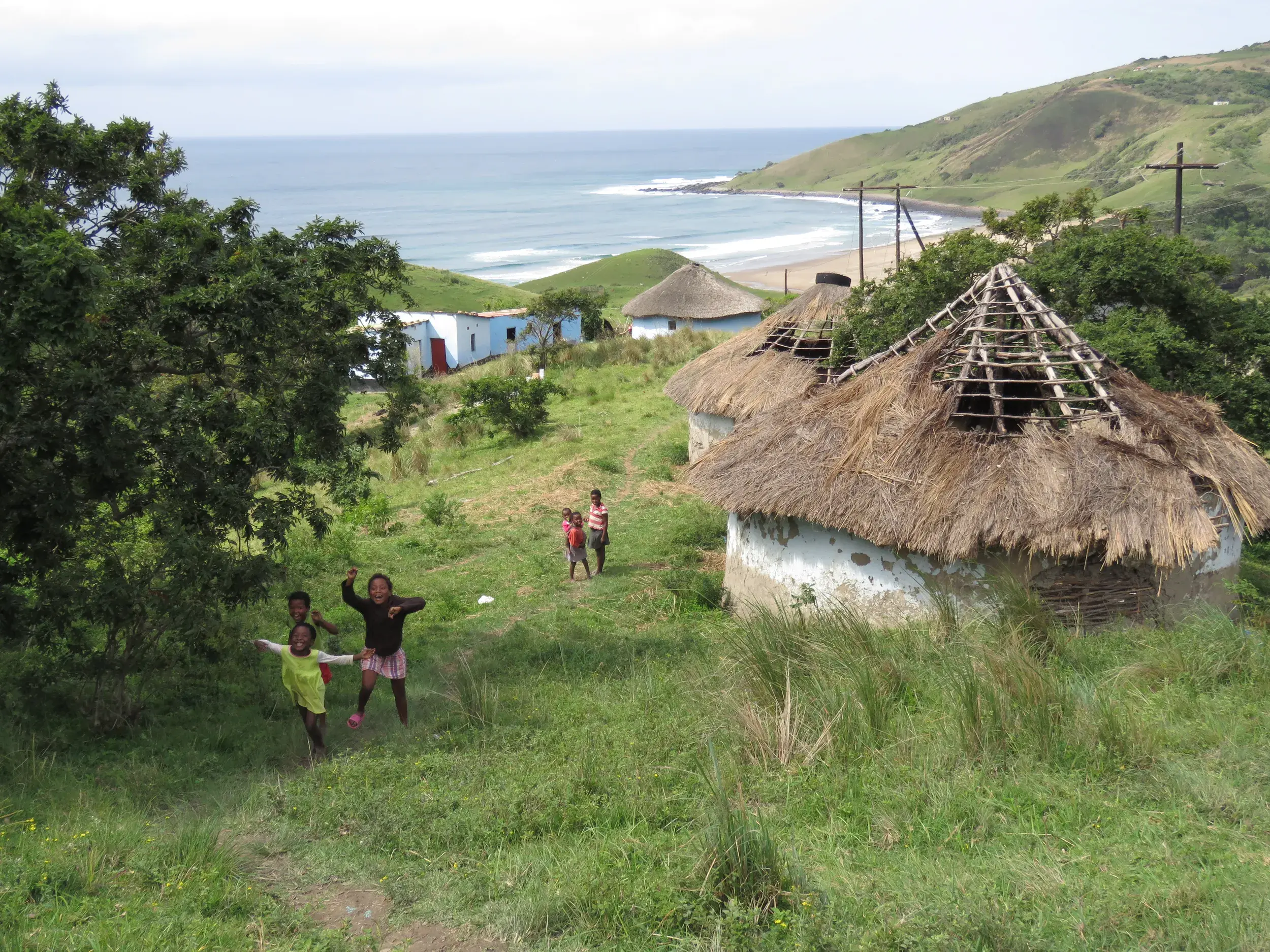 Fröhlich winkende Kinder auf einer Wiese vor traditionellen Rondavels an der Küste der Transkei, der Wild Coast Südafrikas.