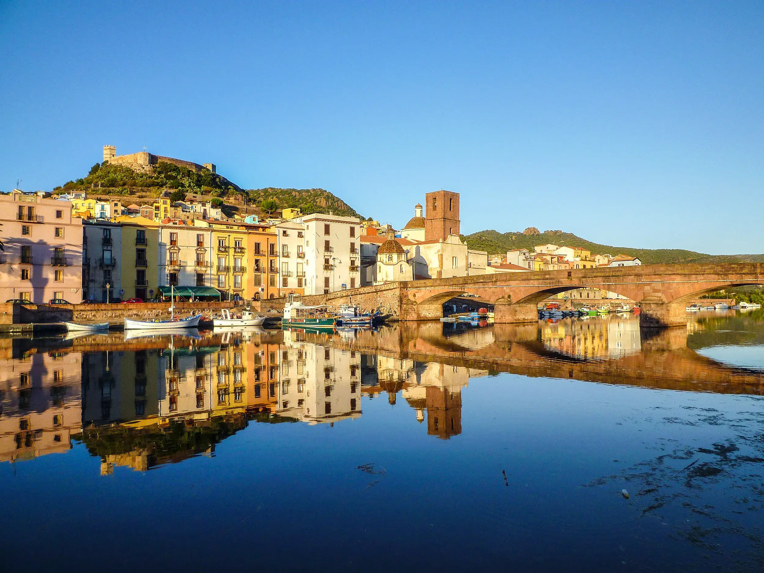 Die bunten Altstadthäuser von Bosa und die alte Steinbrücke spiegeln sich im ruhigen Wasser des Fiume Temo auf Sardinien.