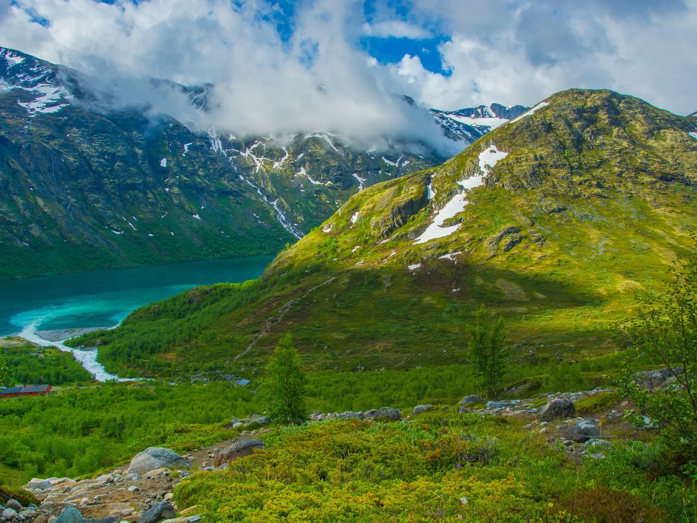Blick auf den türkisen Gjende-See, umgeben von grünen Berghängen mit Schneeresten im Jotunheimen Nationalpark, Norwegen.