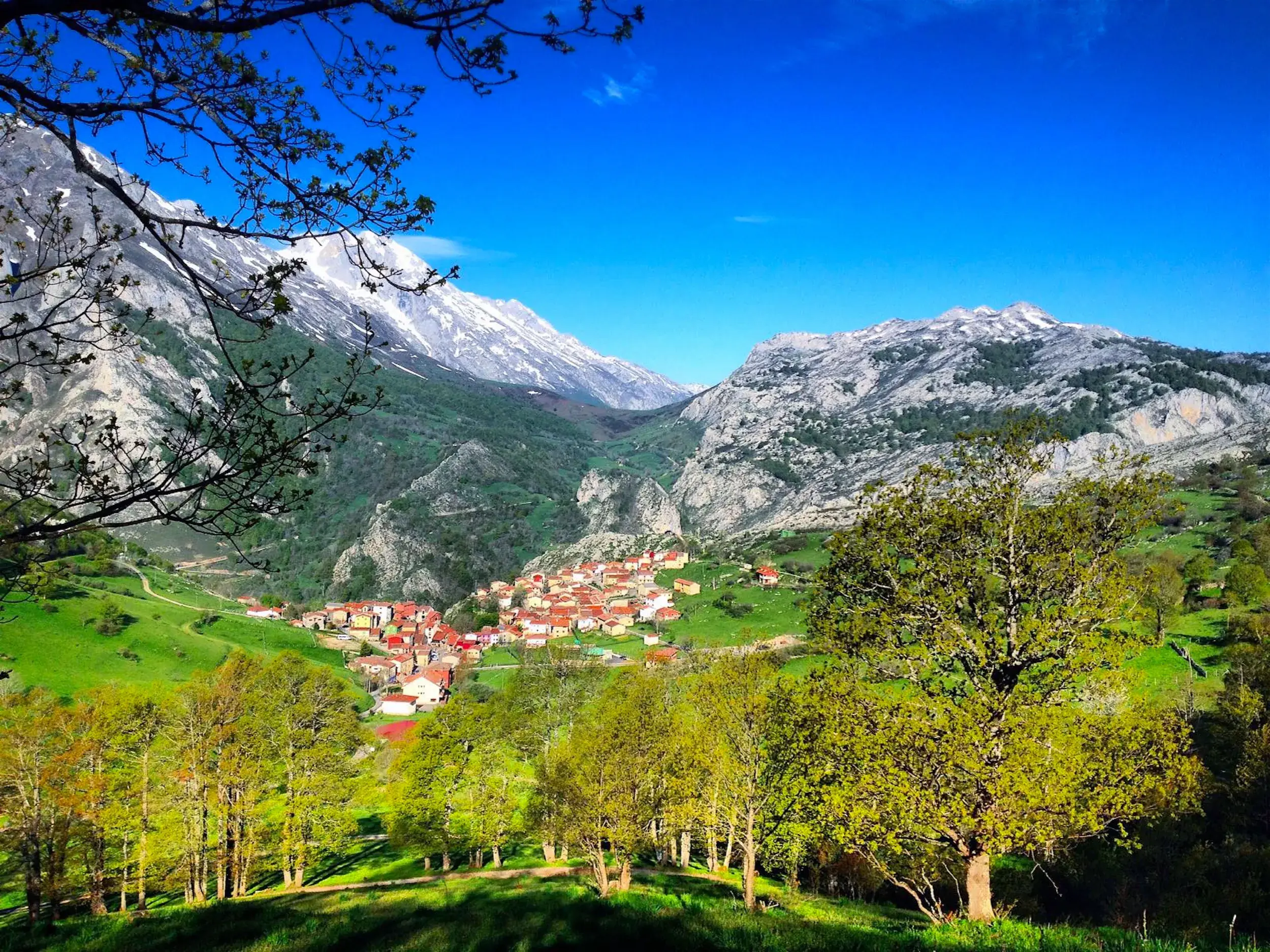 Weitblick auf das Bergdorf Tielves in einem sonnigen, grünen Tal, umgeben von schroffen Gipfeln der Picos de Europa.