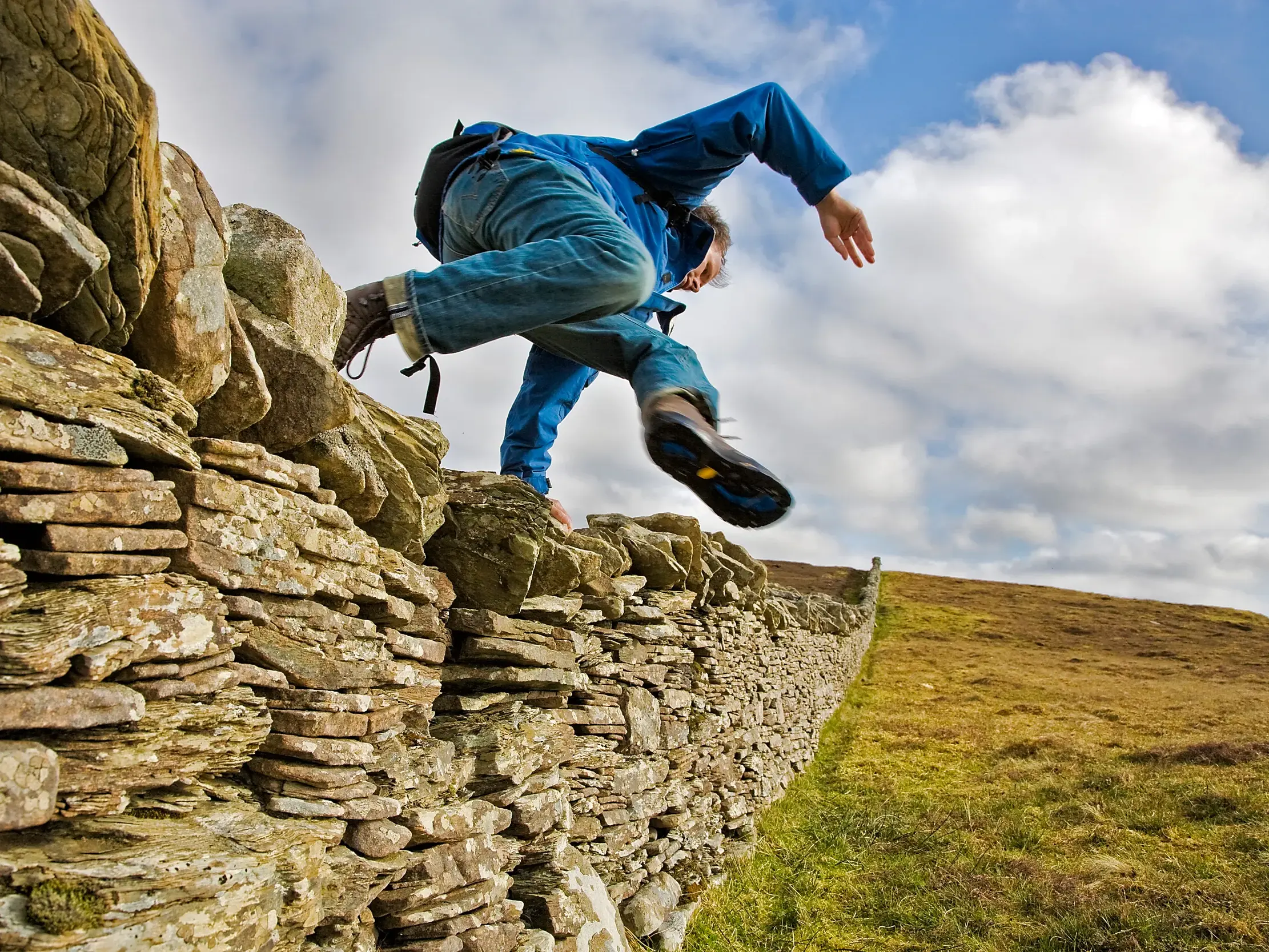 Ein Wanderer springt über eine traditionelle Steinmauer während einer Wanderung in der hügeligen Landschaft der schottischen Highlands.