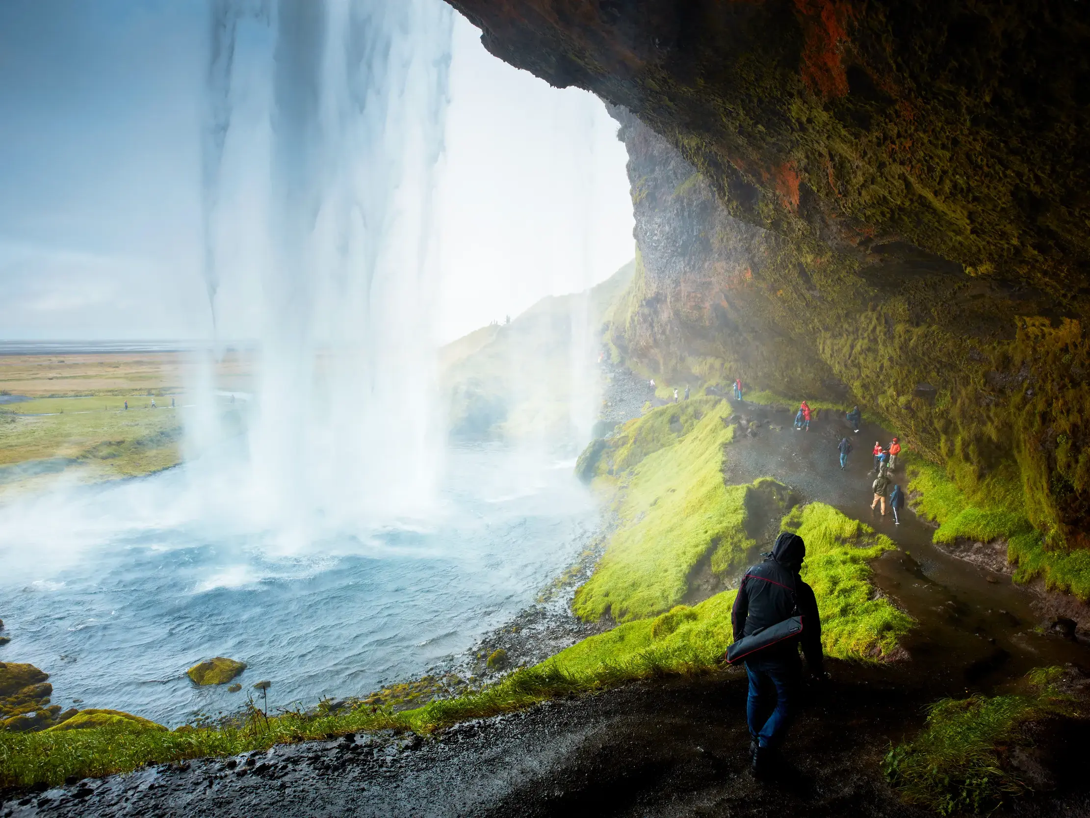Reisende wandern auf einem Pfad hinter dem Wasserschleier des Seljalandsfoss, entlang der moosbewachsenen Felswand in Island.