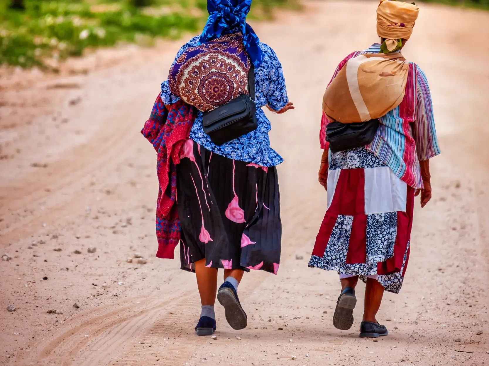 Zwei Frauen in farbenprächtiger, traditioneller Tracht gehen von hinten gesehen auf einer Sandpiste in Namibia.