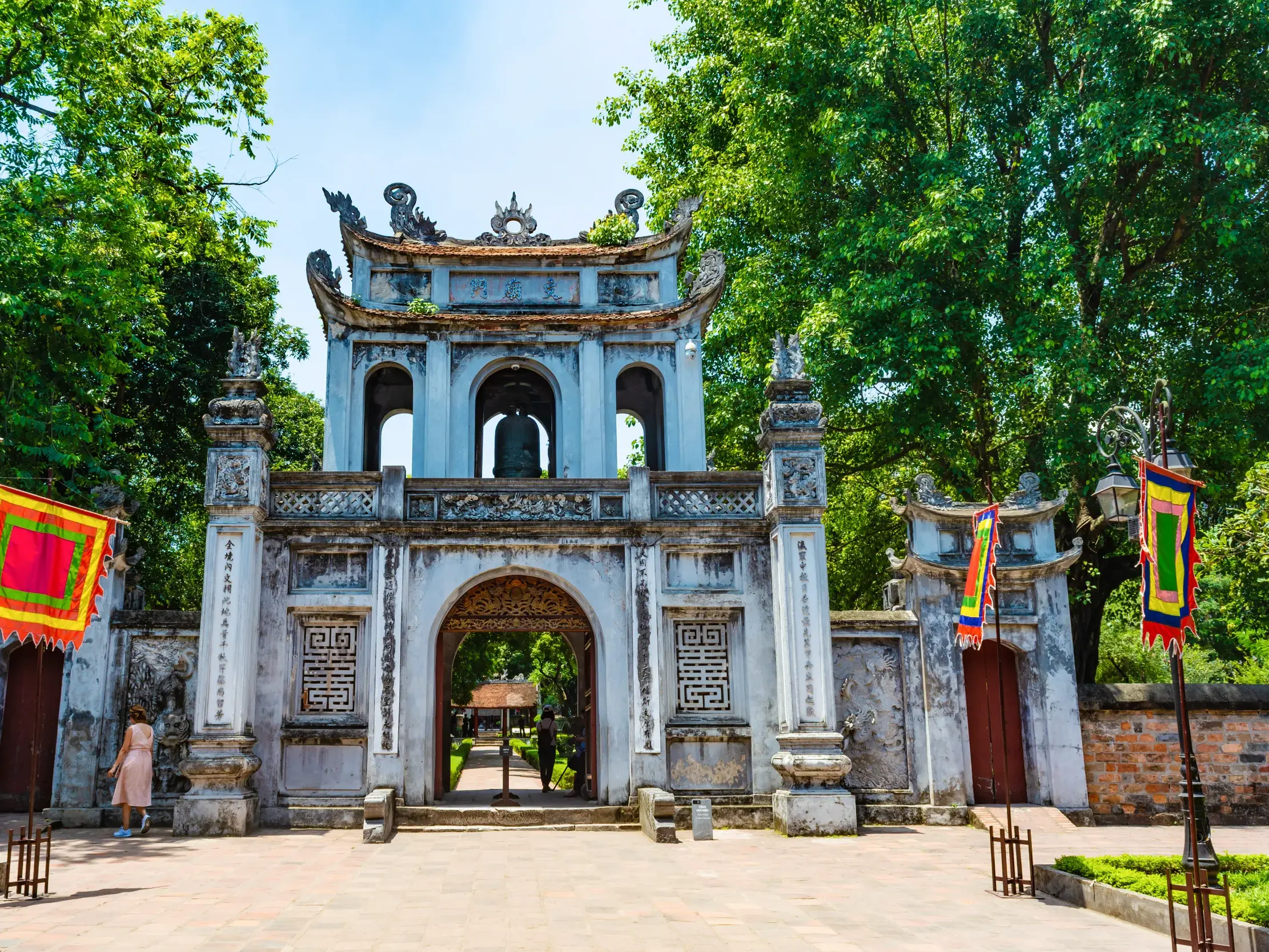 Das große, verzierte Eingangstor des Literaturtempels in Hanoi, flankiert von bunten Fahnen und hohen, grünen Bäumen.