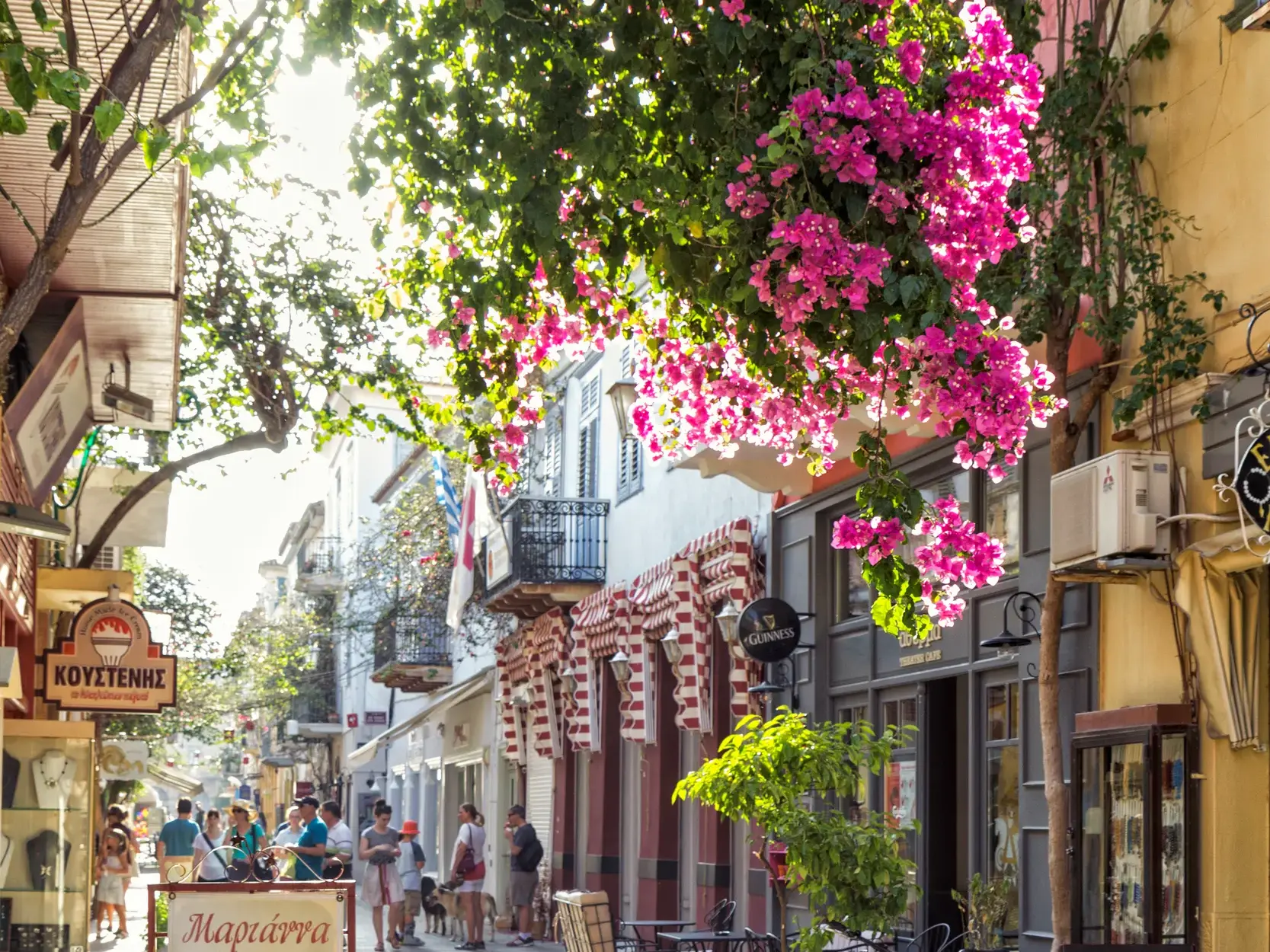 Sonnige Gasse in der malerischen Altstadt von Nauplia, wo pinke Bougainvillea über den Köpfen von Passanten blüht.
