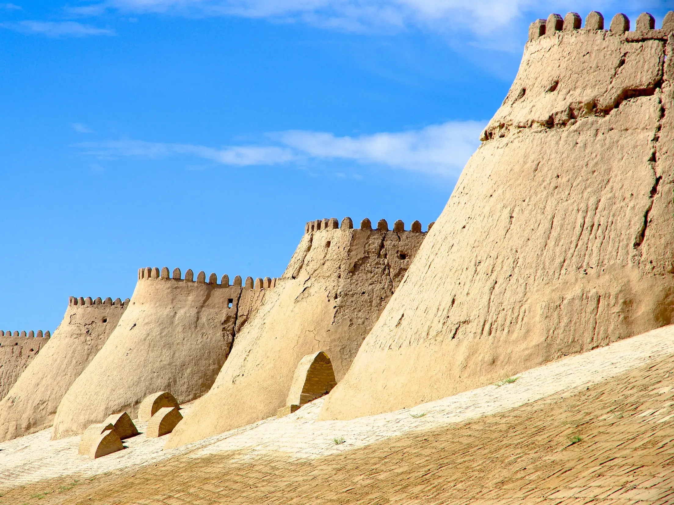 Die mächtige, sandfarbene Lehm-Stadtmauer der historischen Oasenstadt Chiwa in Usbekistan unter strahlend blauem Himmel.