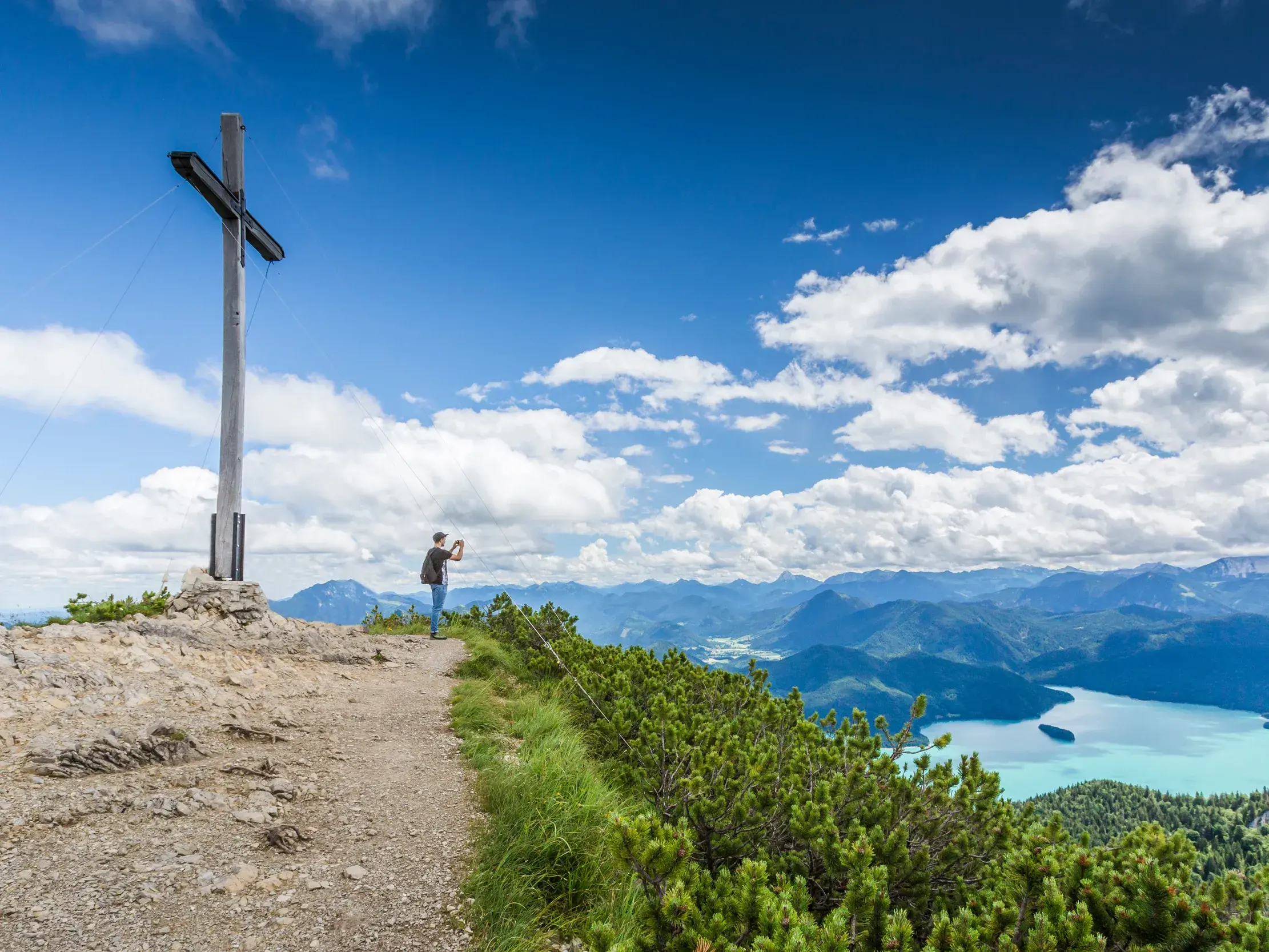 Ein Wanderer am Gipfelkreuz des Herzogstands fotografiert den Panoramablick auf den türkisen Walchensee und das Karwendelgebirge.