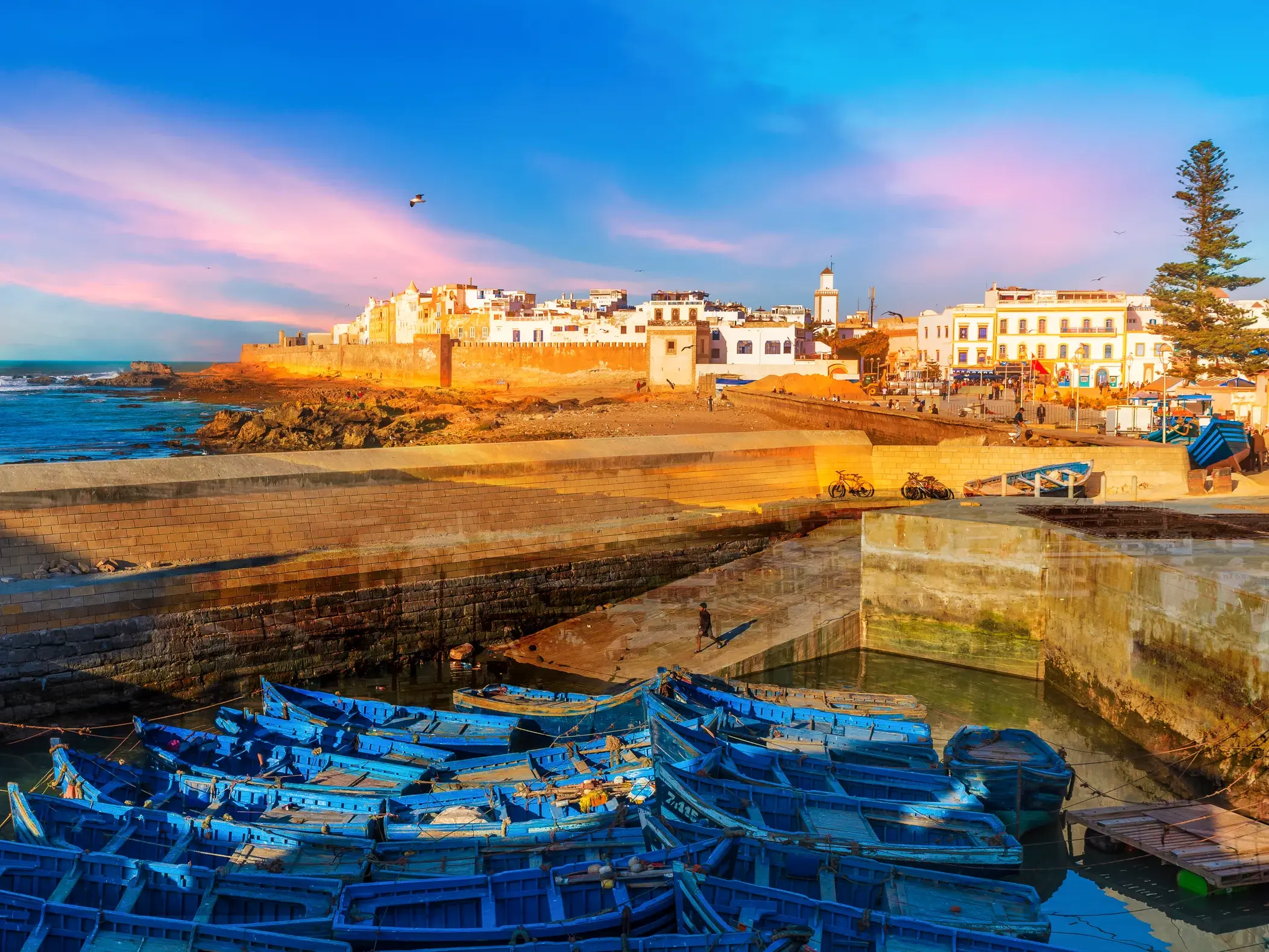 Malerischer Hafen von Essaouira in Marokko mit leuchtend blauen Booten vor der Kulisse der Medina im Abendlicht.