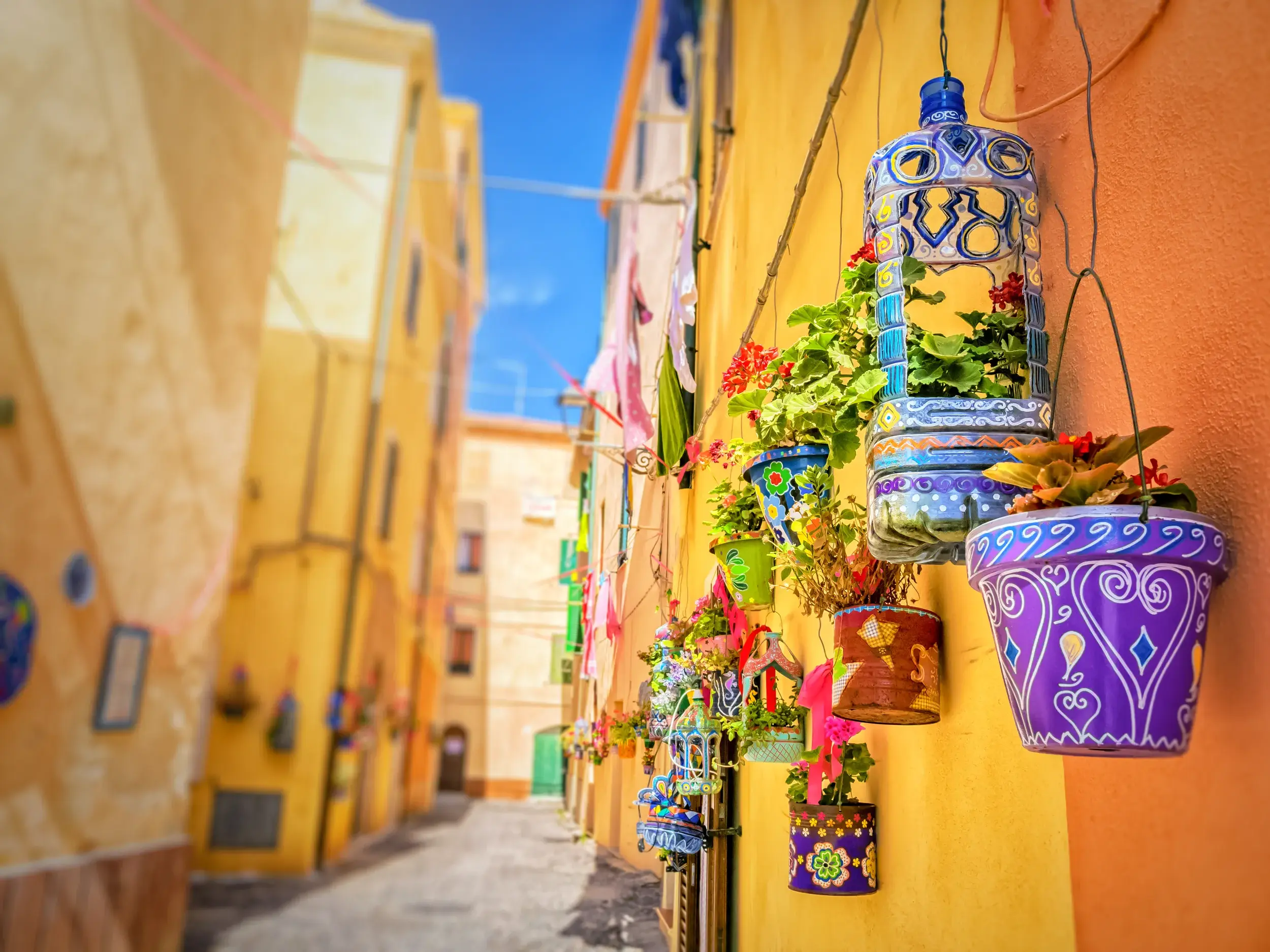 Farbenfrohe, handbemalte Blumentöpfe schmücken eine gelbe Hauswand in einer sonnigen Gasse der Altstadt von Bosa auf Sardinien.