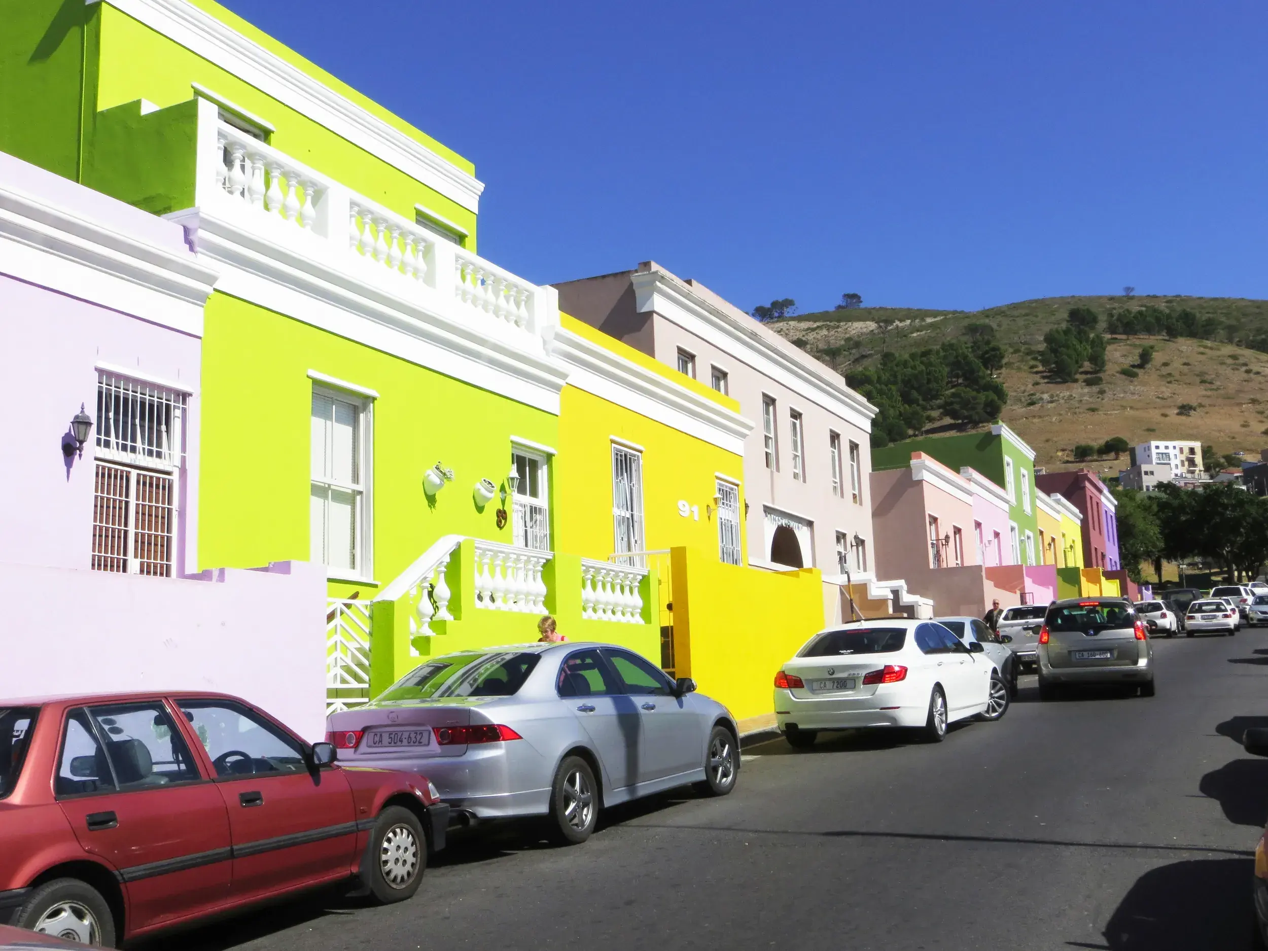 Farbenfrohe Häuserfassaden im Bo-Kaap Viertel in Kapstadt säumen eine Straße, im Hintergrund ein grüner Hügel.