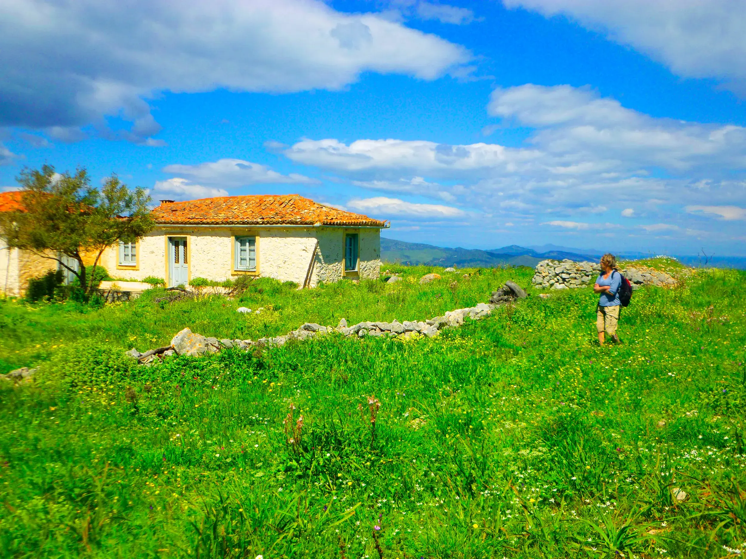 Eine Wanderin steht auf einer Blumenwiese vor einem Steinhaus auf der griechischen Insel Hydra mit Blick auf den Saronischen Golf.